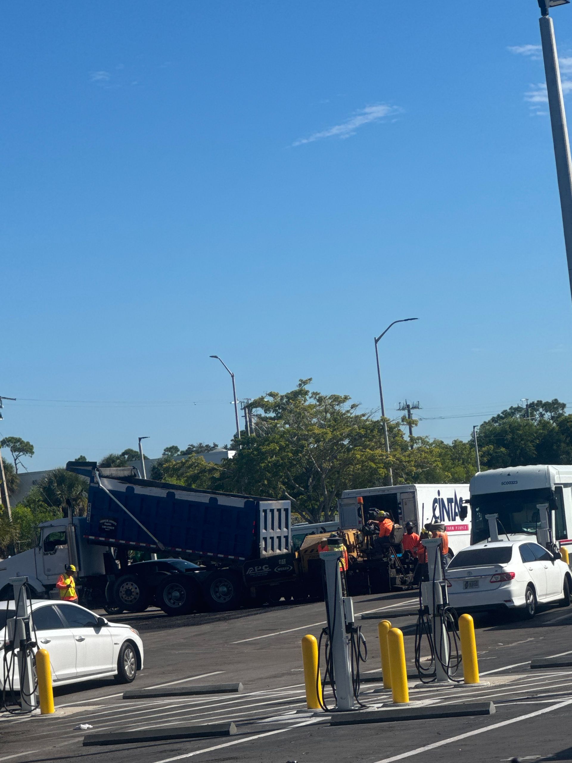 Dump truck unloading asphalt on a road, construction workers in vests, cars, and a blue sky.
