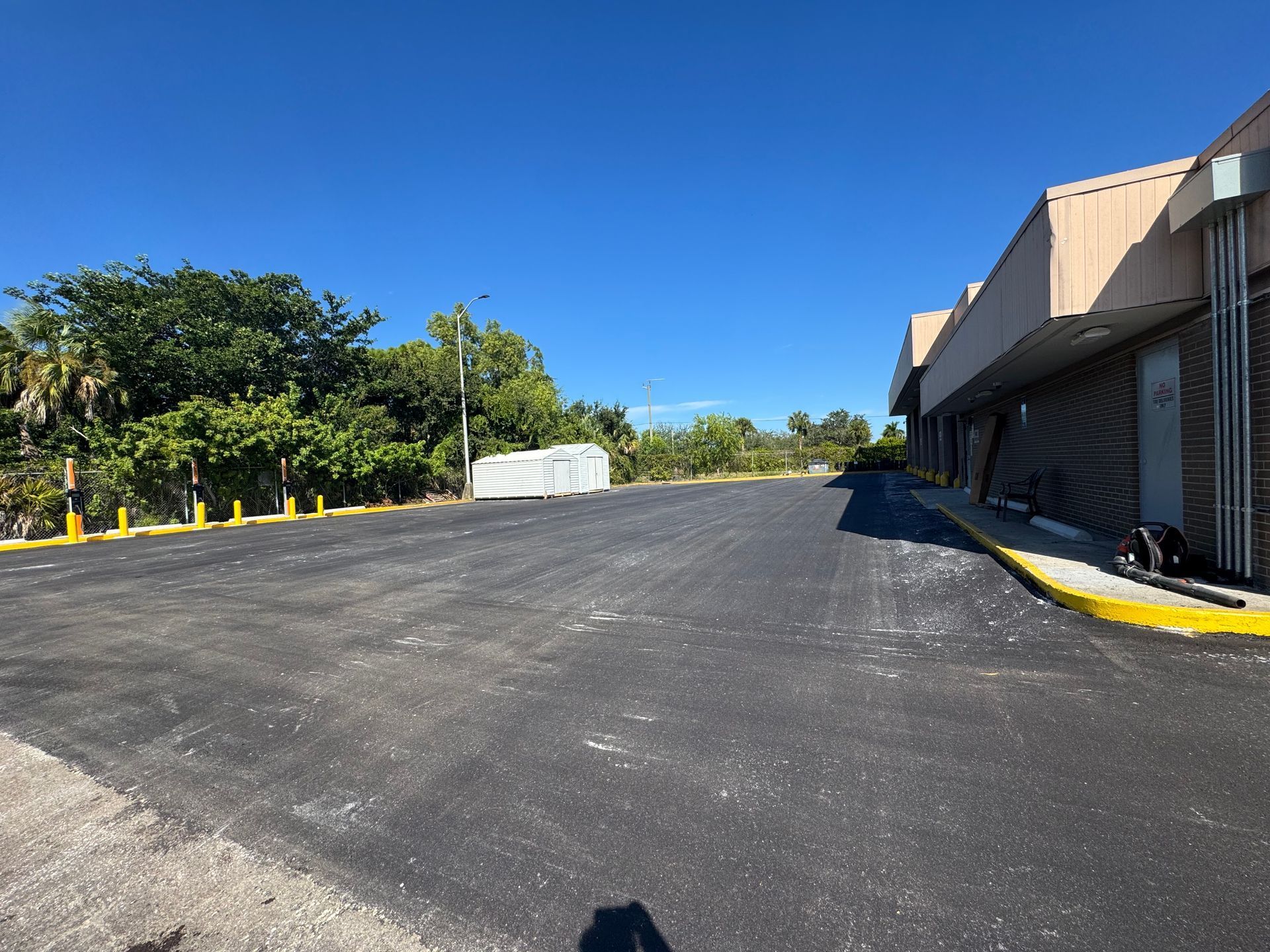 Paved parking area next to a commercial building and greenery, under a clear, blue sky.