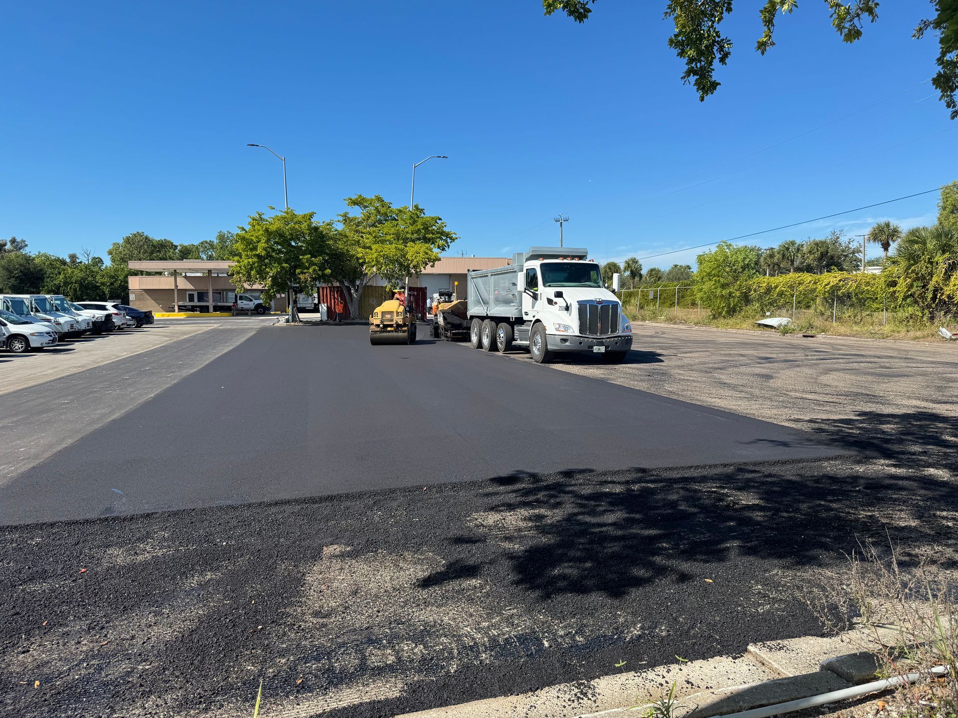 Asphalt paving of a parking lot. Dump truck, roller, workers, and a building are in the scene under a blue sky.
