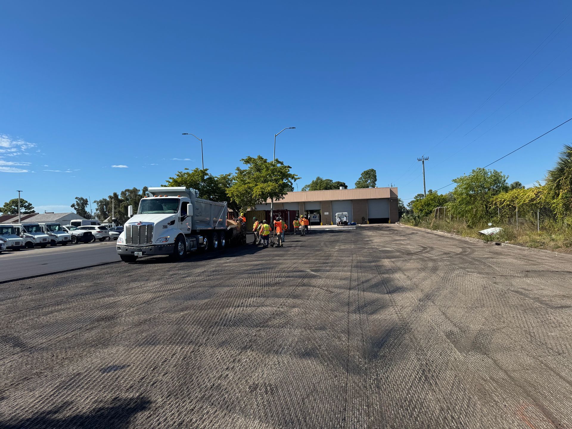 Dump truck unloading asphalt on a paved lot in front of a small building under a blue sky.