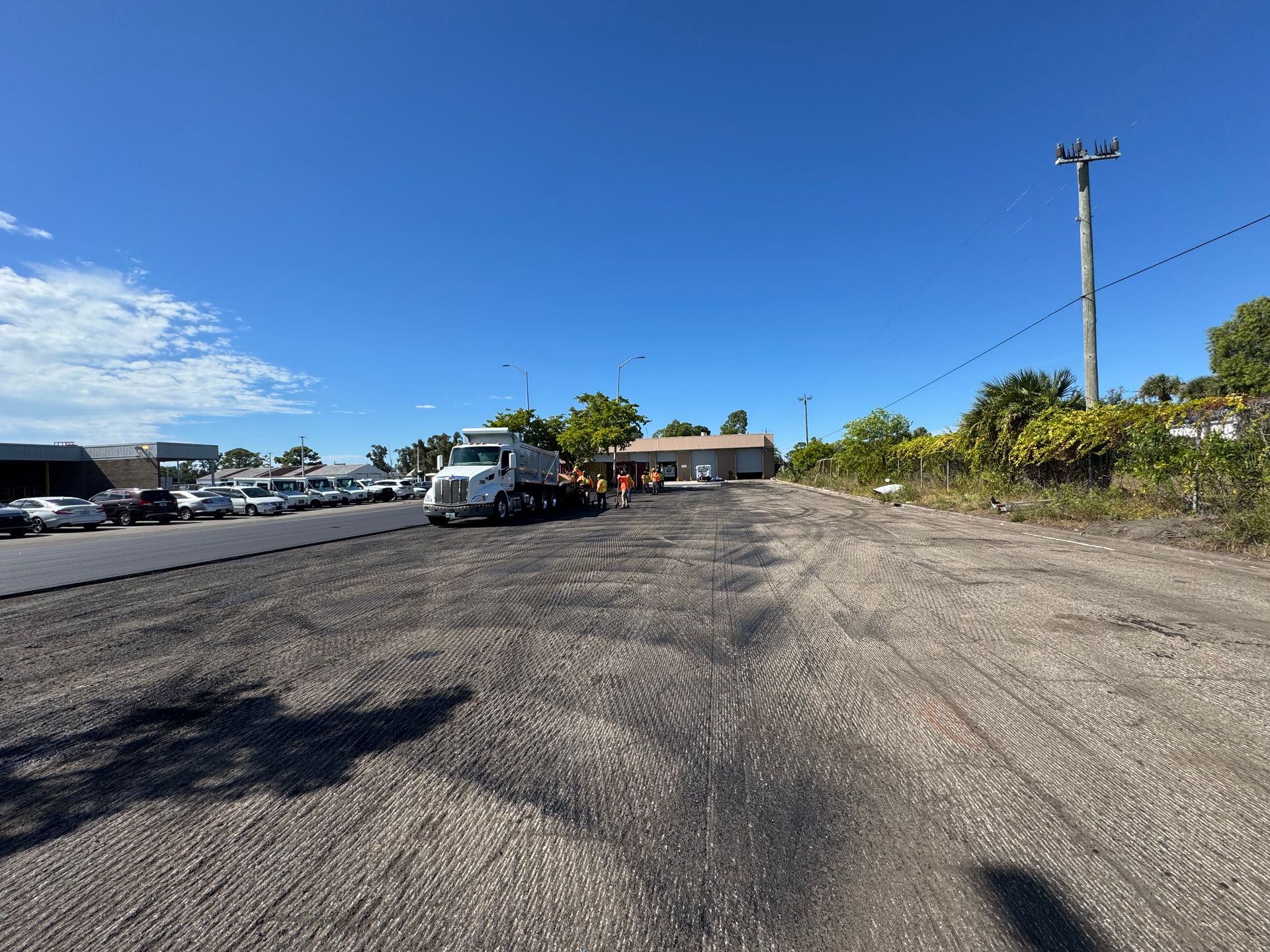 Truck parked on a gravel surface next to an asphalt road, a line of cars, and a building under a blue sky.