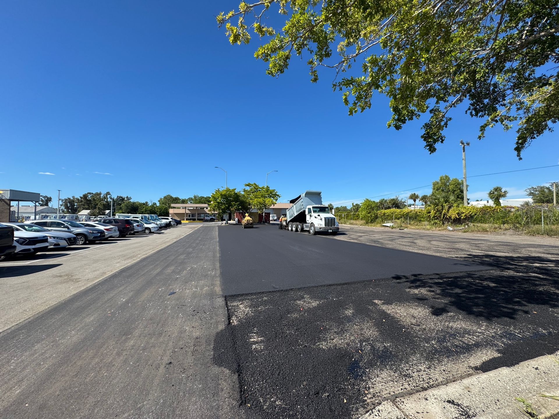 Asphalt paving of a parking area by a truck; blue sky, cars parked nearby.
