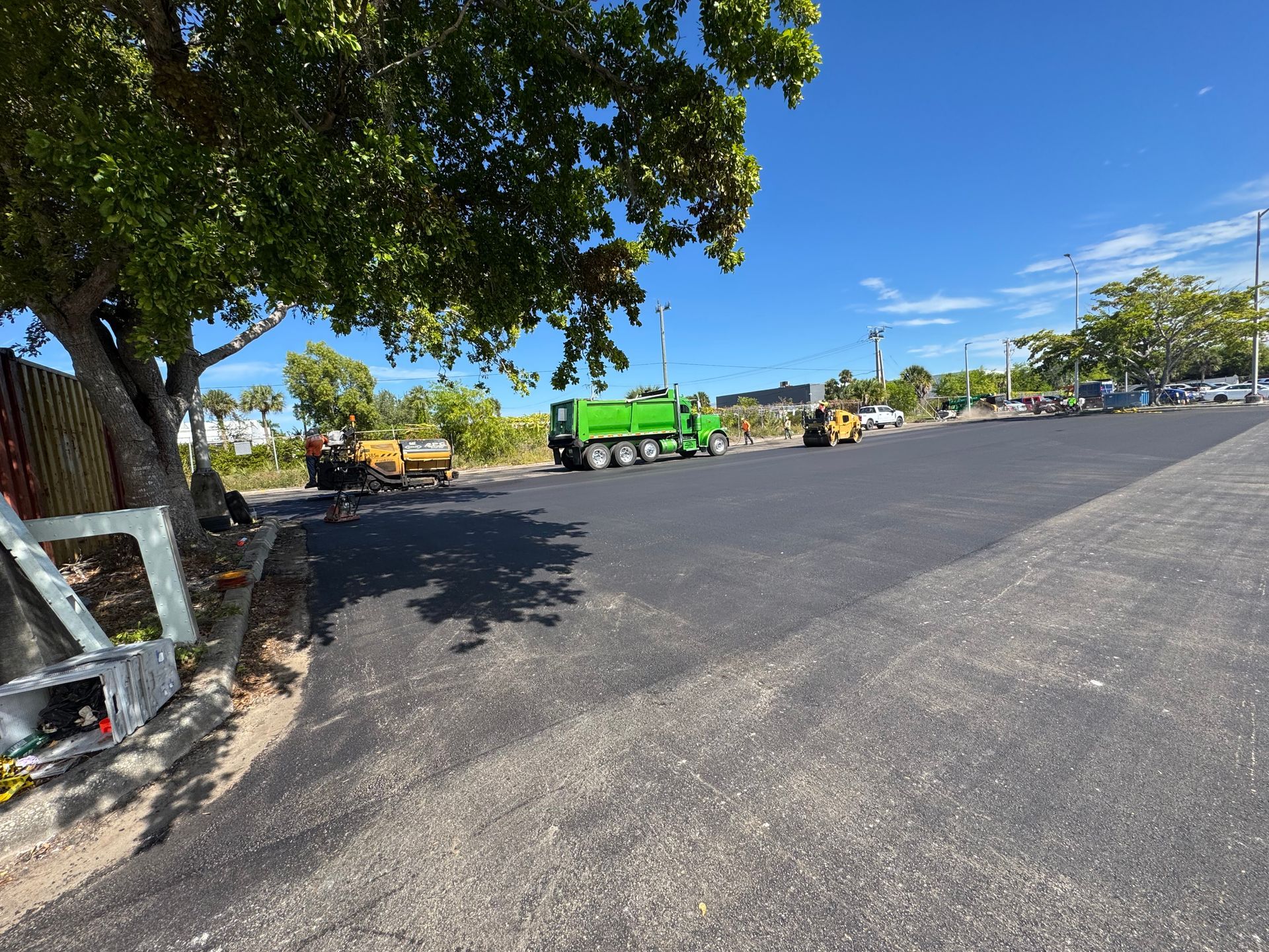 Newly paved asphalt parking area with construction equipment and a green dump truck under a blue sky.