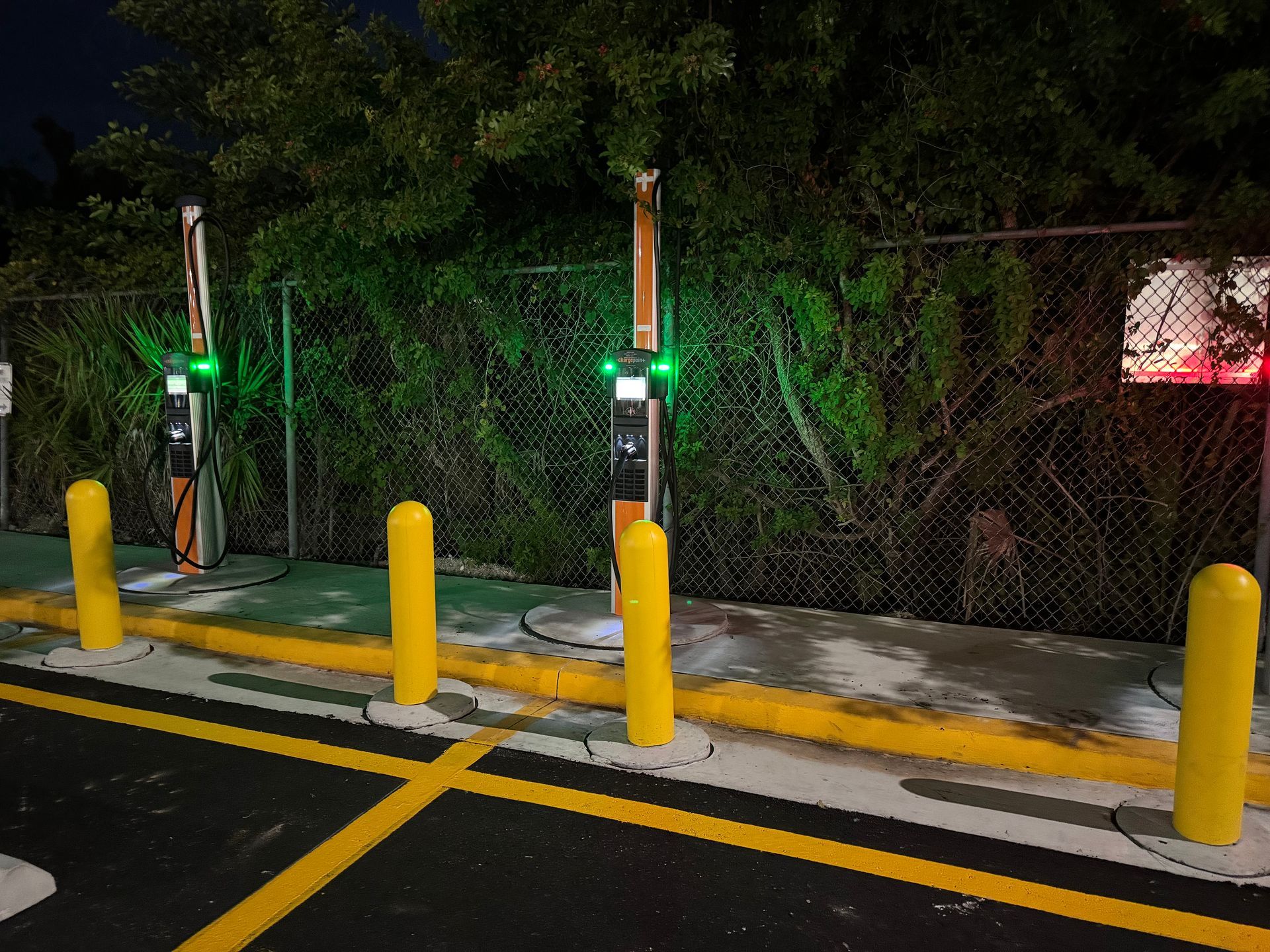 Electric vehicle chargers at night with green lights. Yellow bollards and fence are visible.