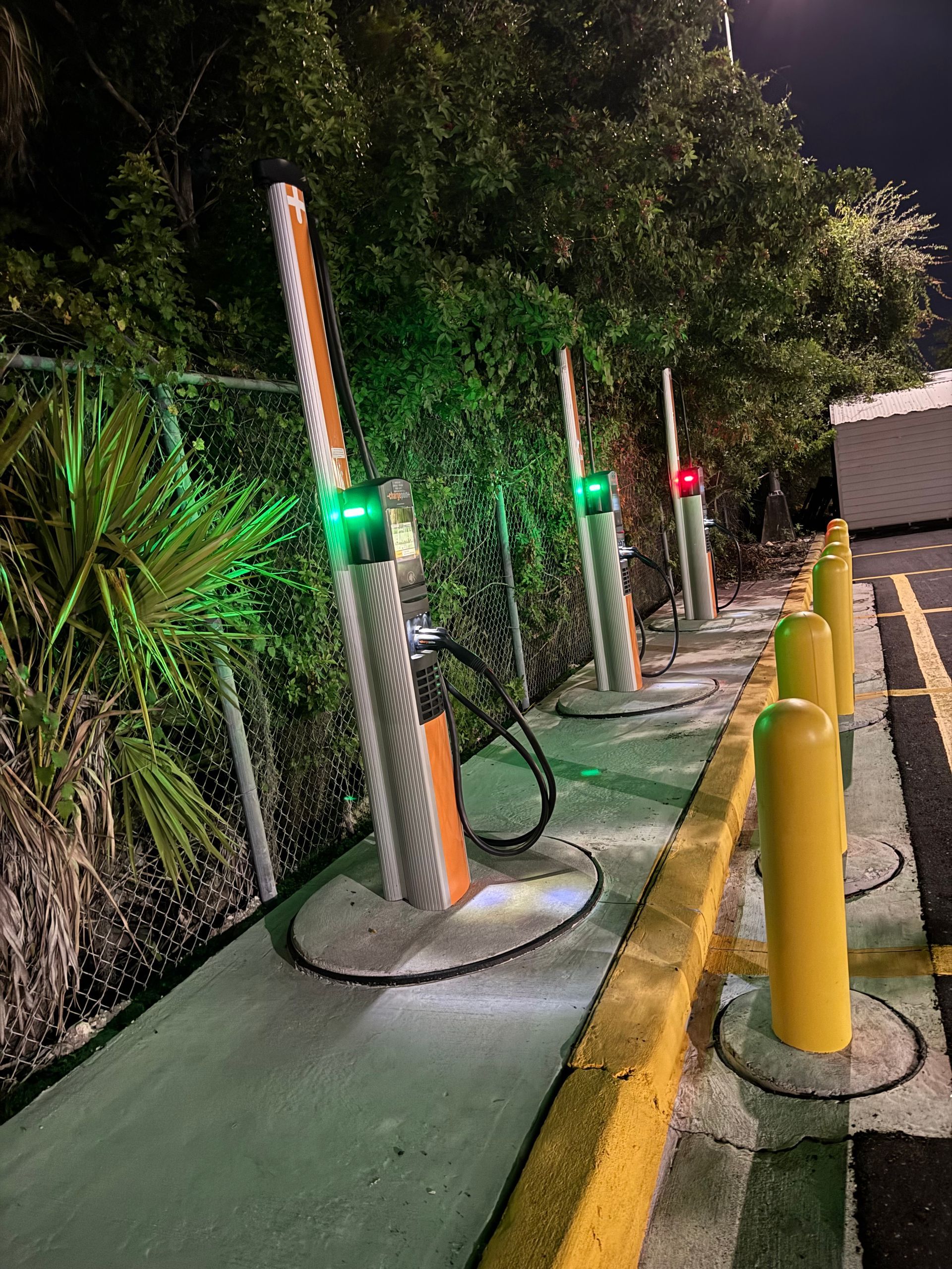 Electric vehicle chargers at night with green and red lights, next to a fence.