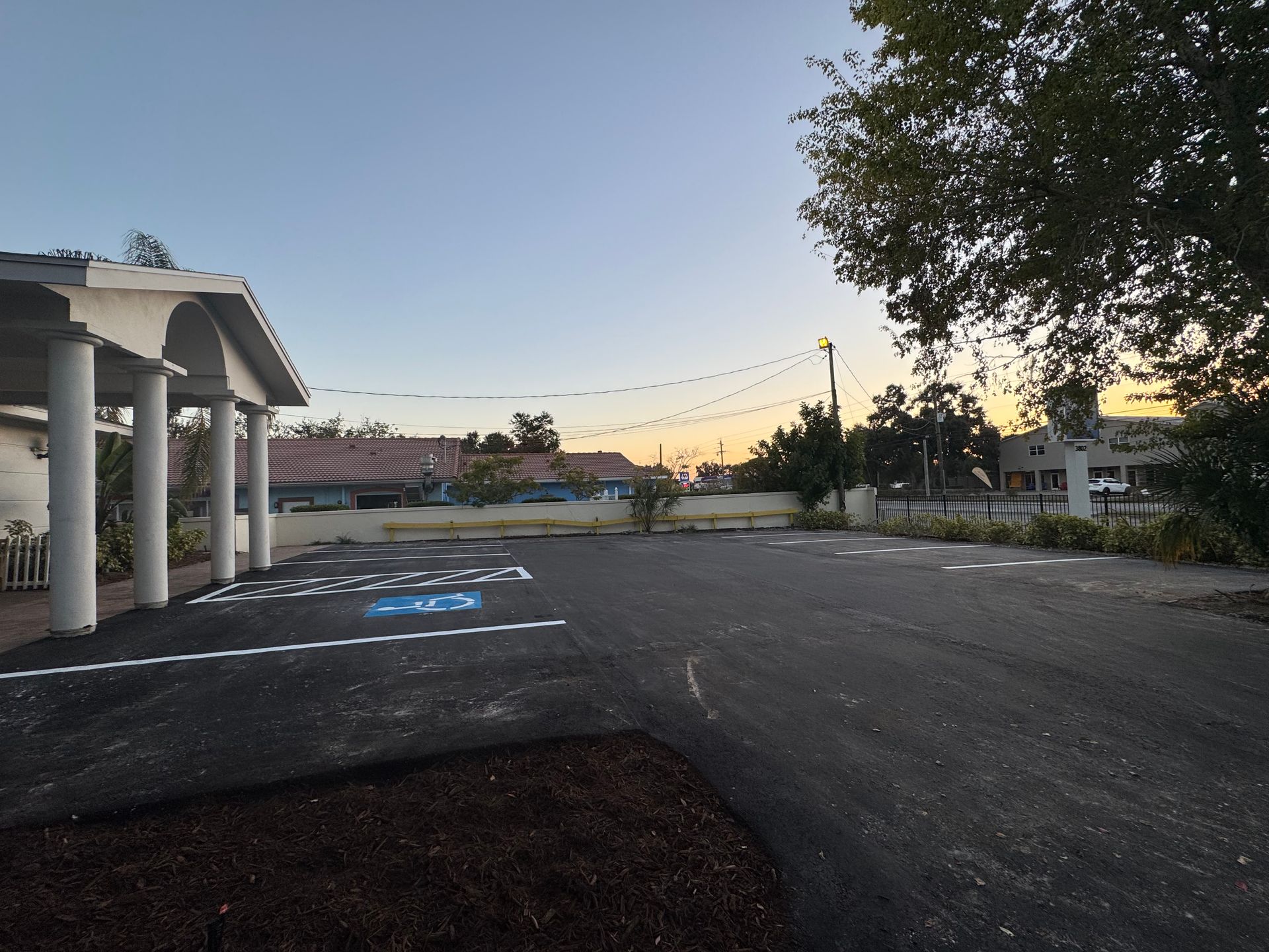 Paved parking lot with designated spaces, a building with columns, and a tree, under a dusky sky.