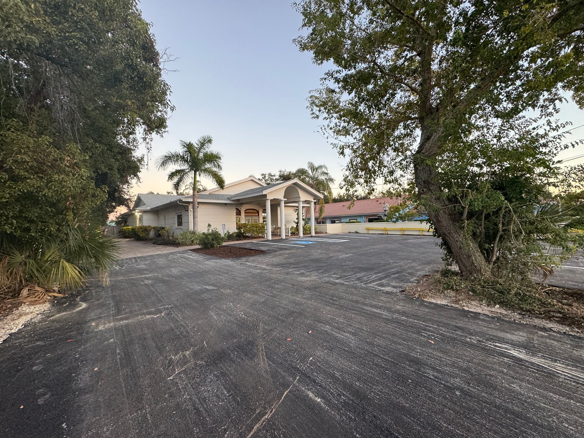 A single-story building with a paved driveway, flanked by trees and greenery under a clear sky.