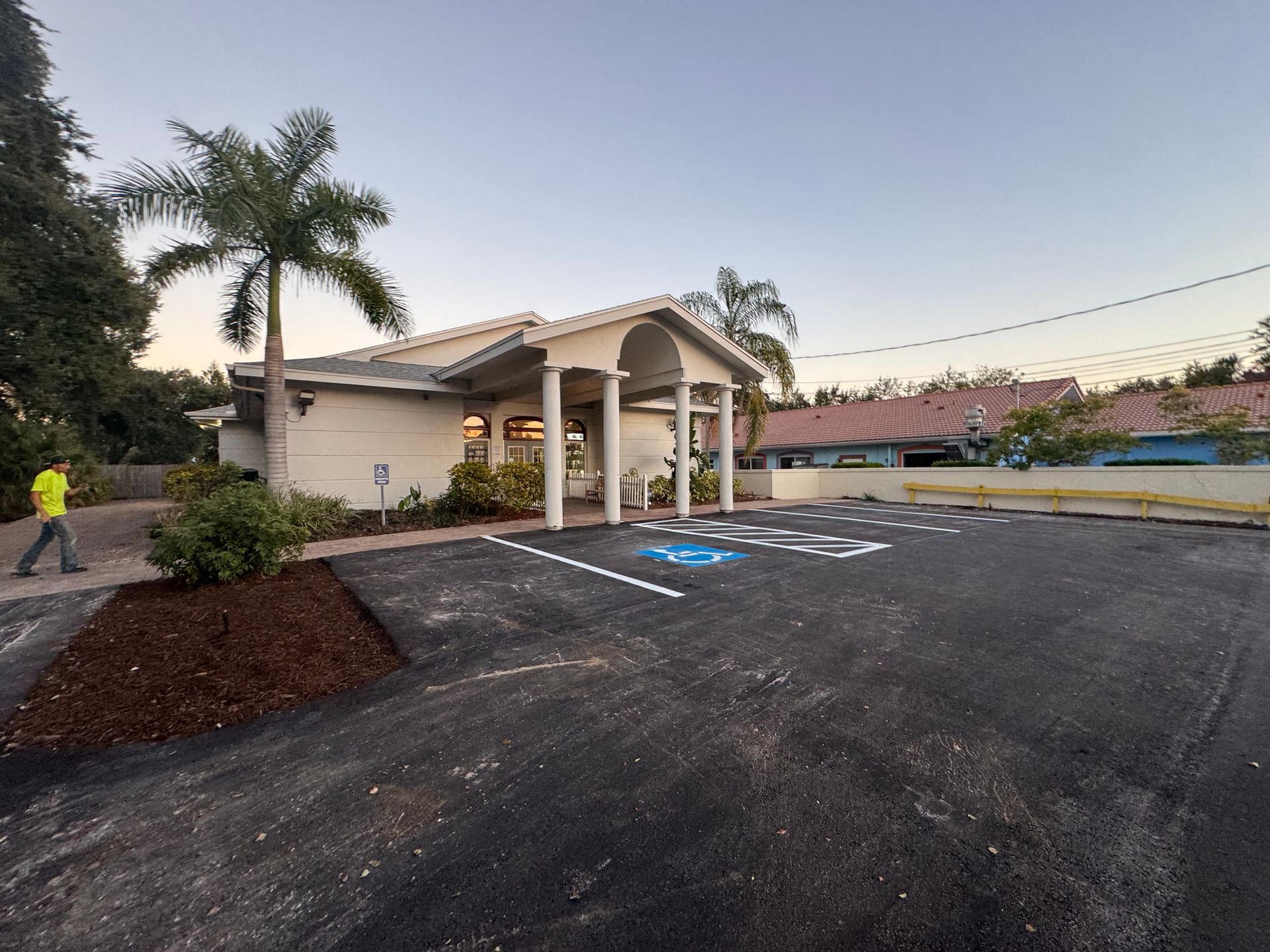 Exterior view of a cream-colored building with a covered entrance, palm trees, and a fresh black asphalt parking lot.