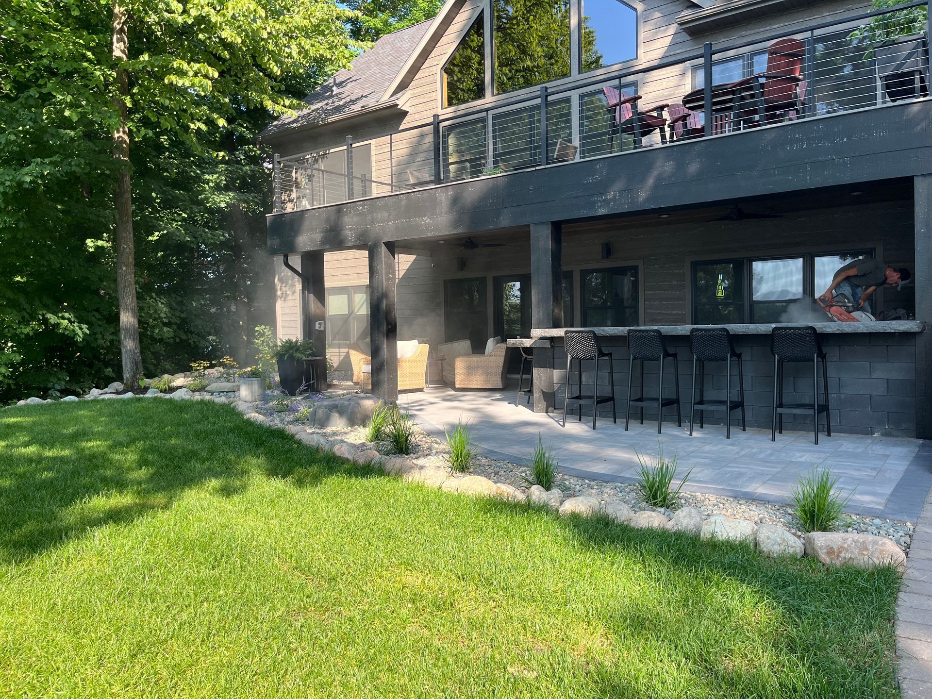 Modern home with outdoor bar, deck, and green lawn. Grey brick, glass railing, and black bar stools.