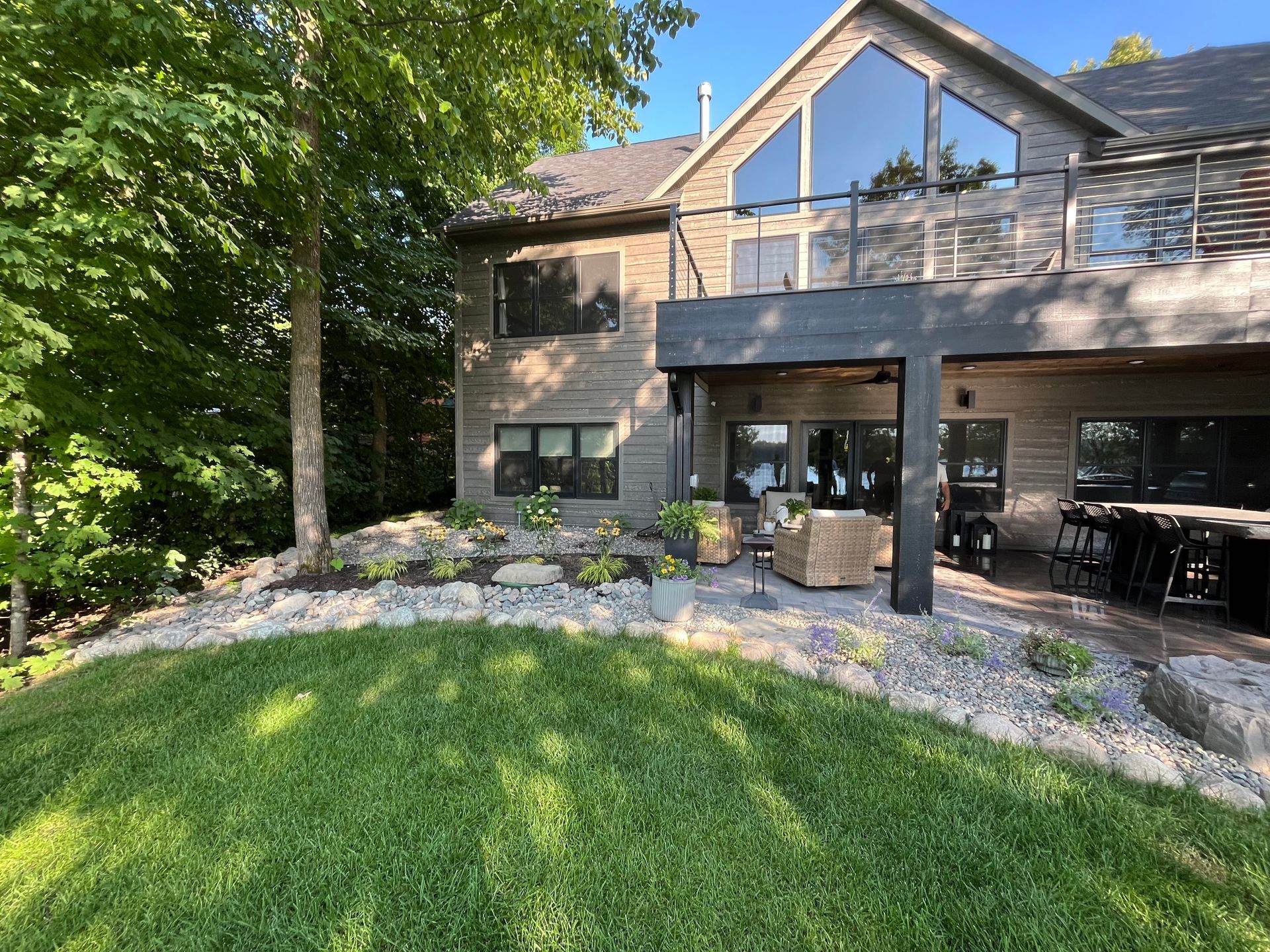 Lakeside house with large windows, deck, and patio. Green lawn, trees, and rocky landscaping.