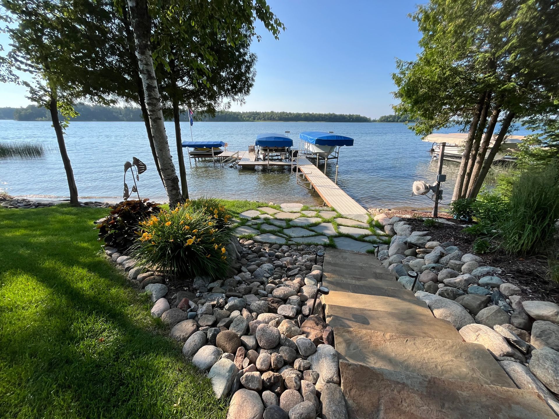 Stone path leads to a dock with blue awnings on a lake, surrounded by trees and grass.