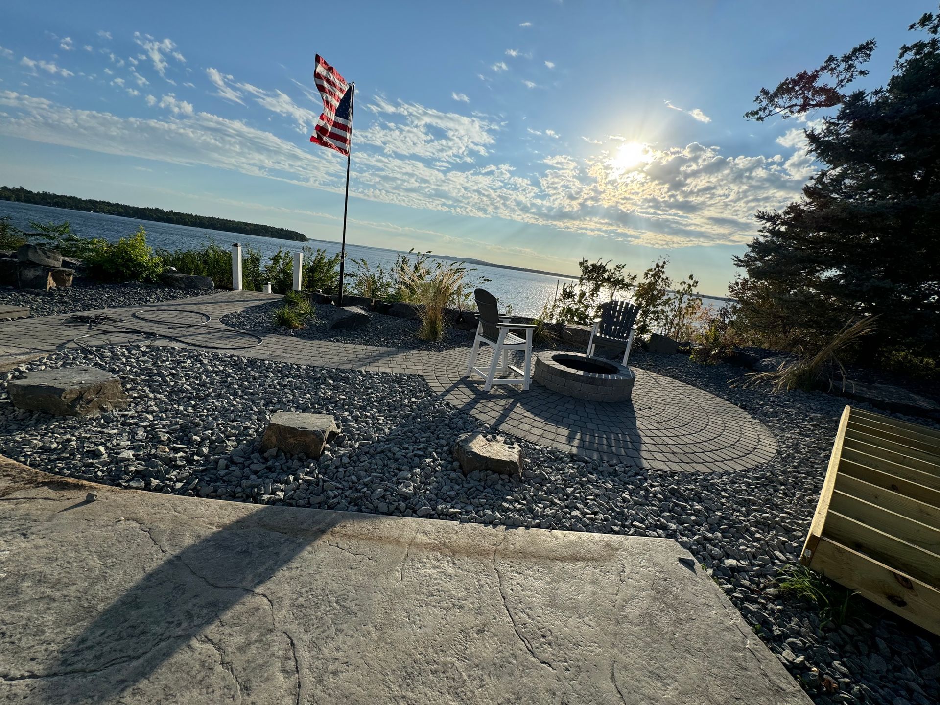 A waterfront patio with a fire pit, American flag, and water view under a sunny sky.
