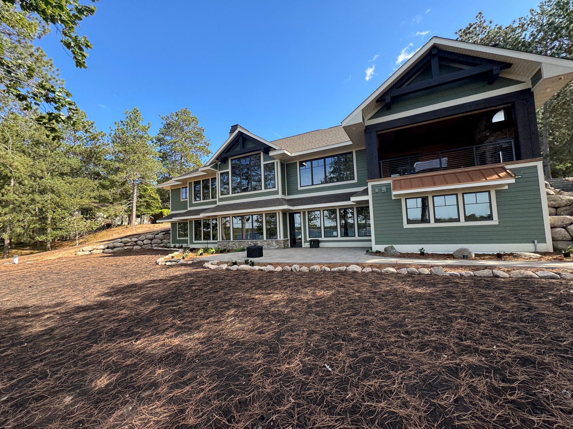Green lake house with large windows, set in a natural landscape under a blue sky.
