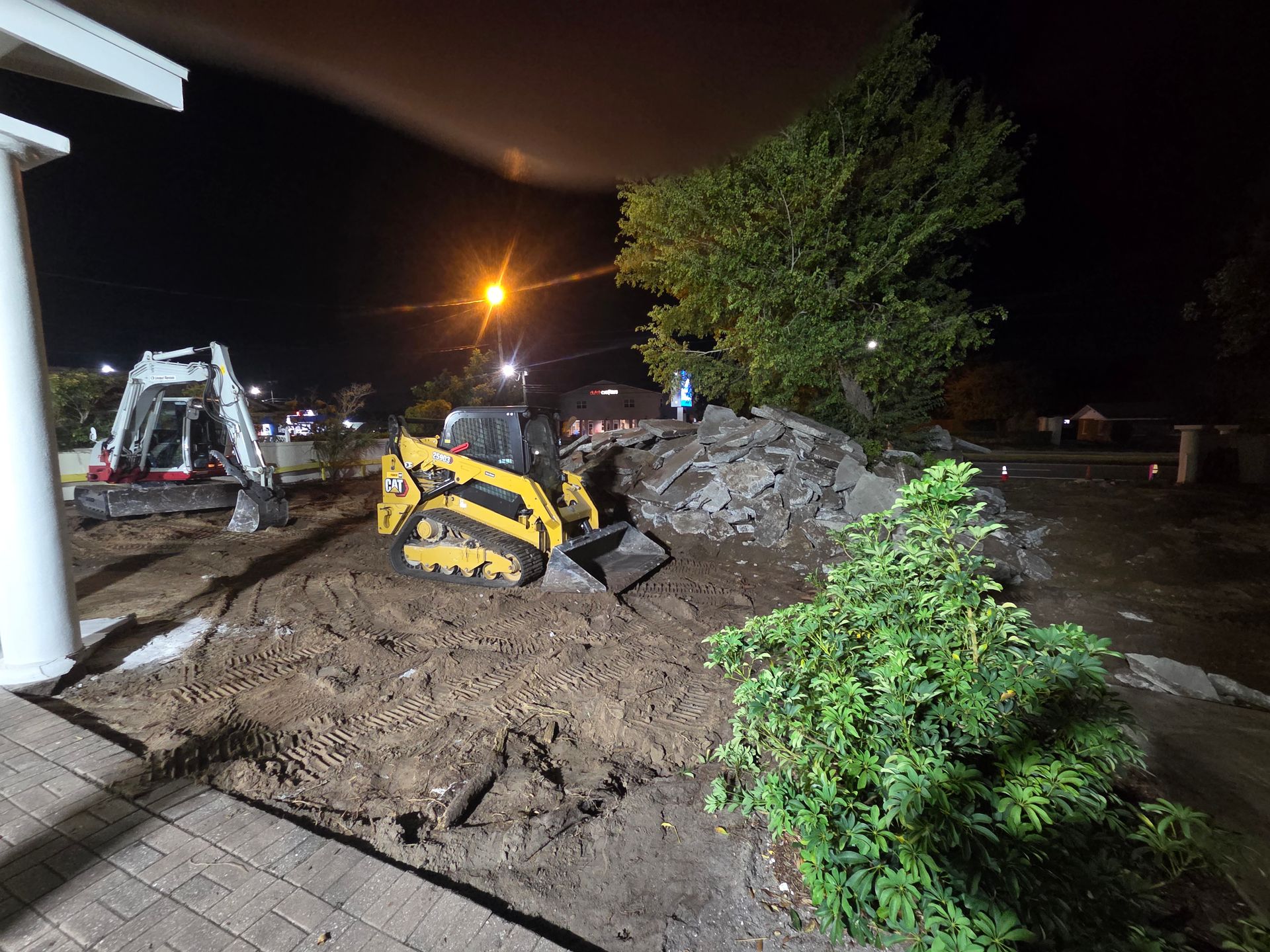 Construction site at night with a skid steer, excavator, pile of debris, and a bush. Dark setting.