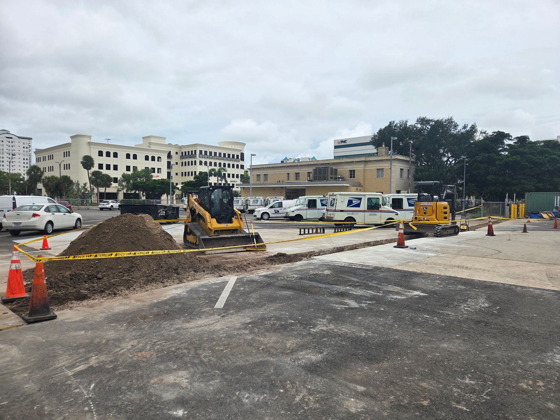 Construction zone with heavy machinery, piles of dirt, and barriers in a parking area, with buildings in the background.