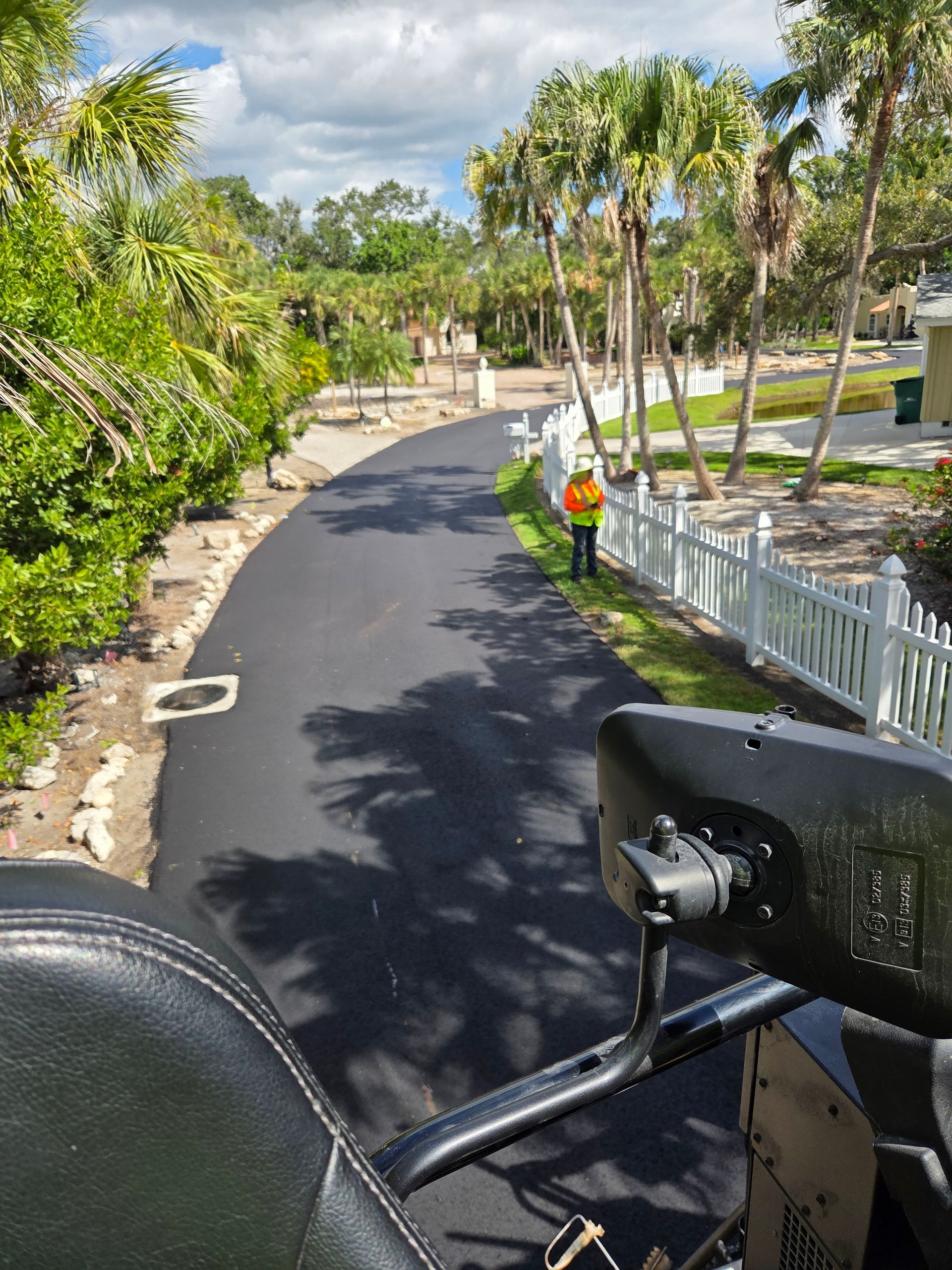 Newly paved road with a worker wearing a safety vest; lined with palm trees and white picket fence.