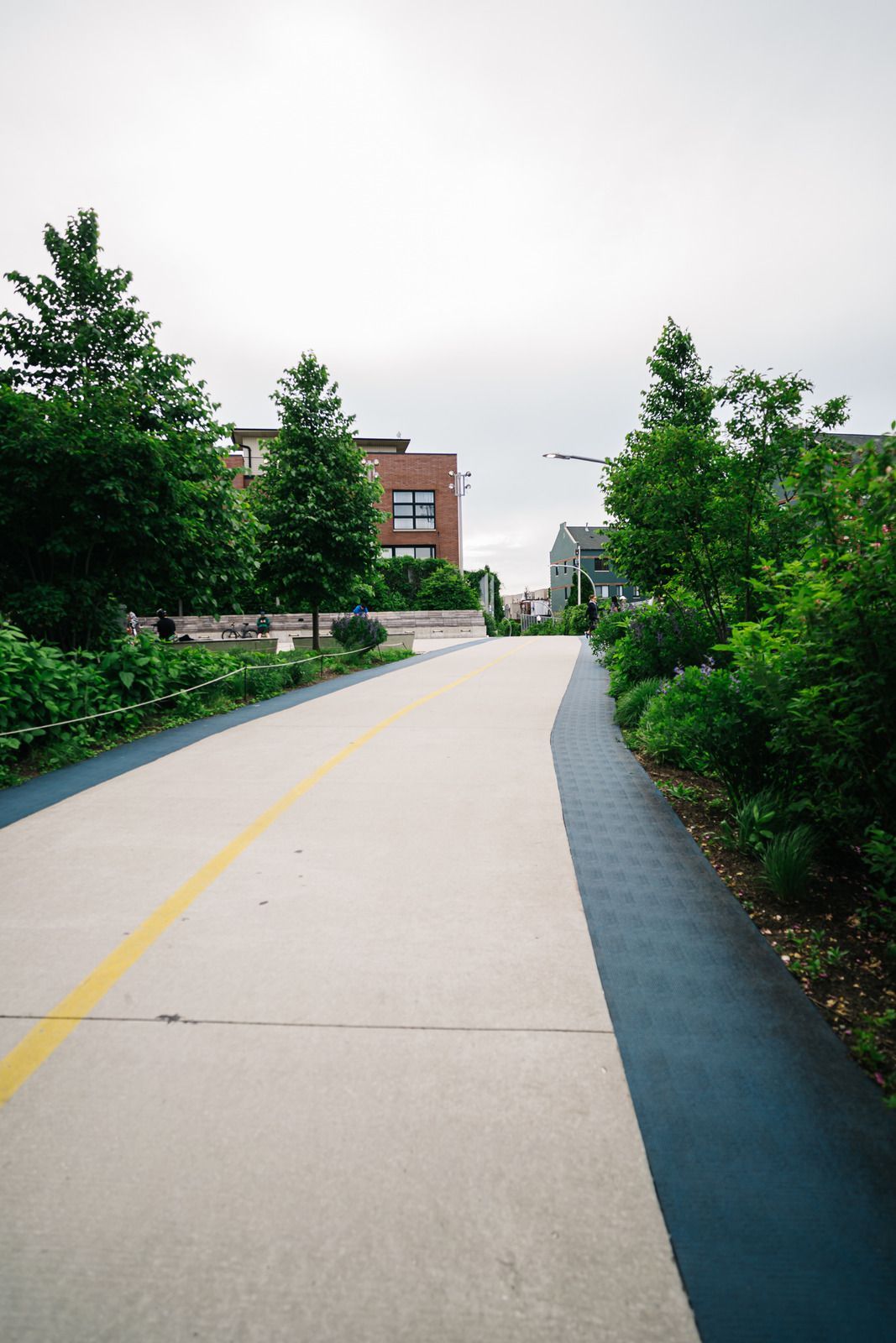 Paved path with a yellow line, flanked by greenery and buildings under a cloudy sky.