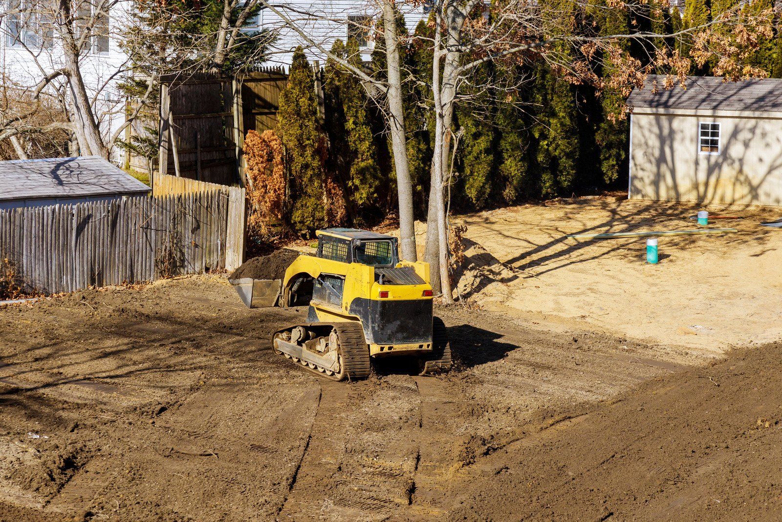 Yellow track loader on a dirt lot, preparing a yard for construction, with trees and a shed in the background.