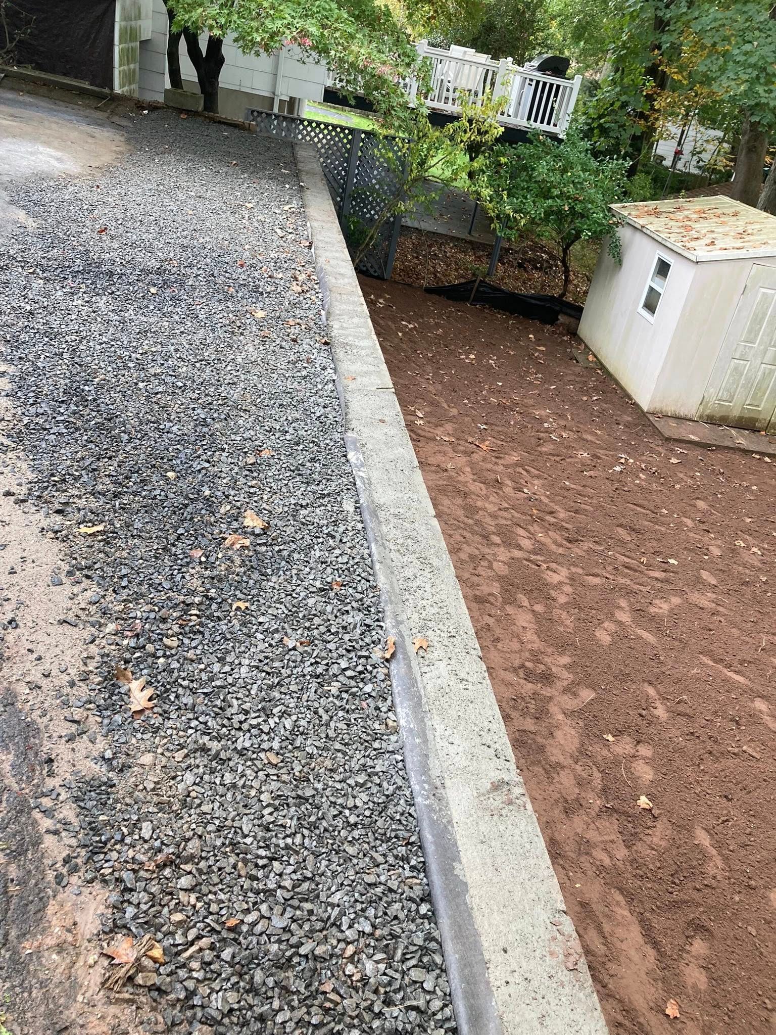 Gravel driveway next to a concrete border, leading to a dirt area with a small white shed.