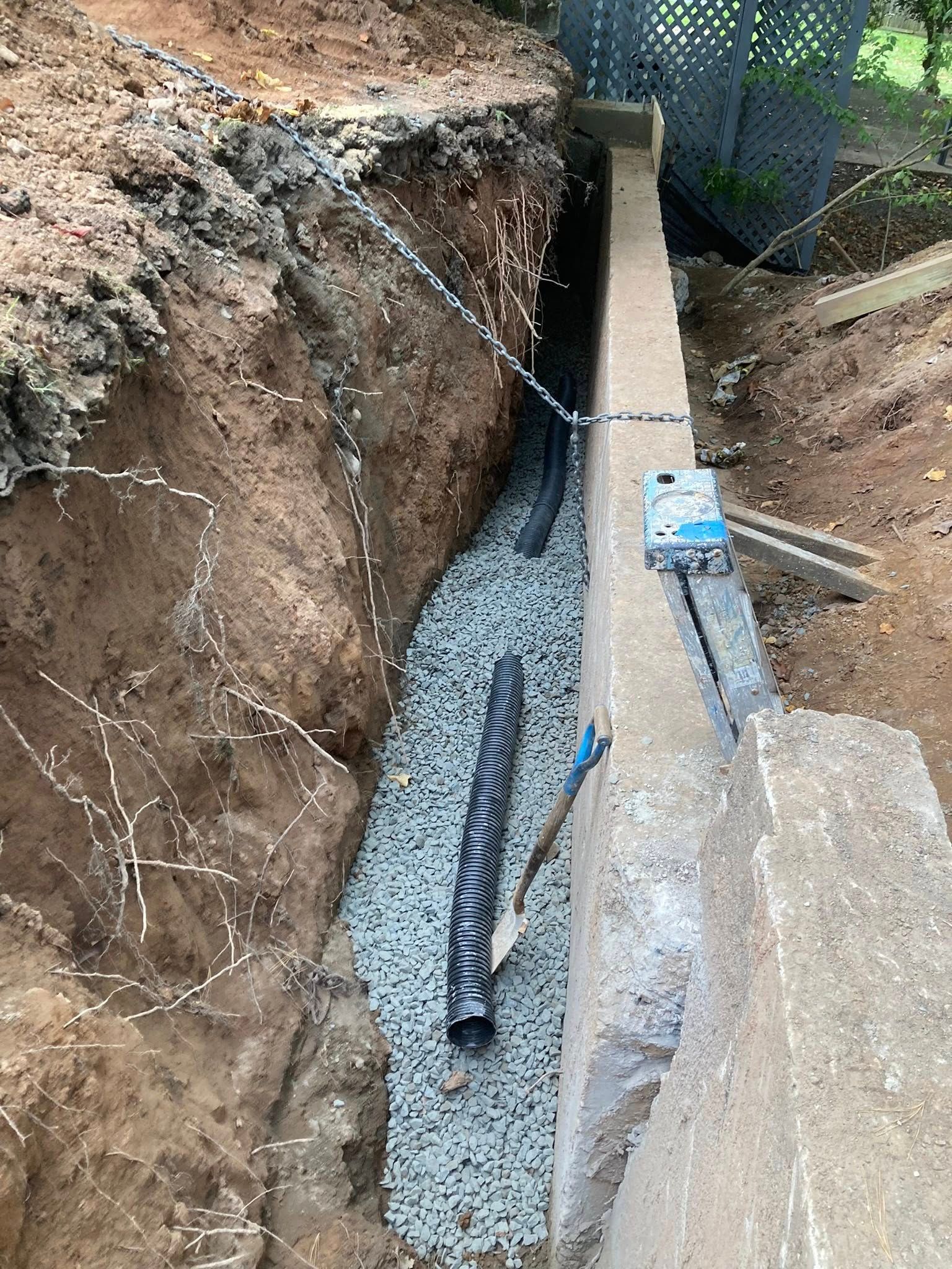 Trench with drainage pipe and gravel alongside a concrete wall. Earth bank on the left and a fence in the background.