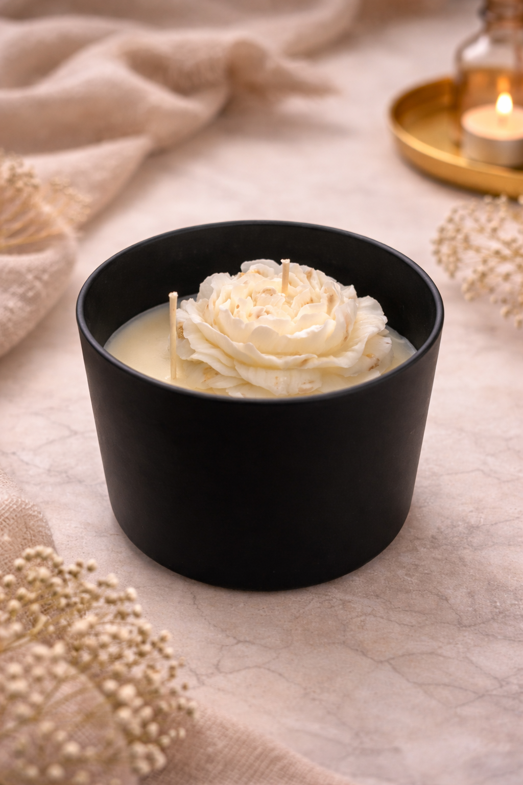 Een zwarte kaars in een pot met een wit bloemmotief, naast een brandende kaars en gedroogde bloemen.