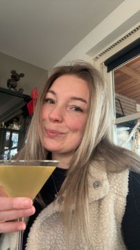 Een persoon met lang blond haar, die een martiniglas vasthoudt, glimlacht lichtjes en draagt ​​een crèmekleurig, pluizig vest.