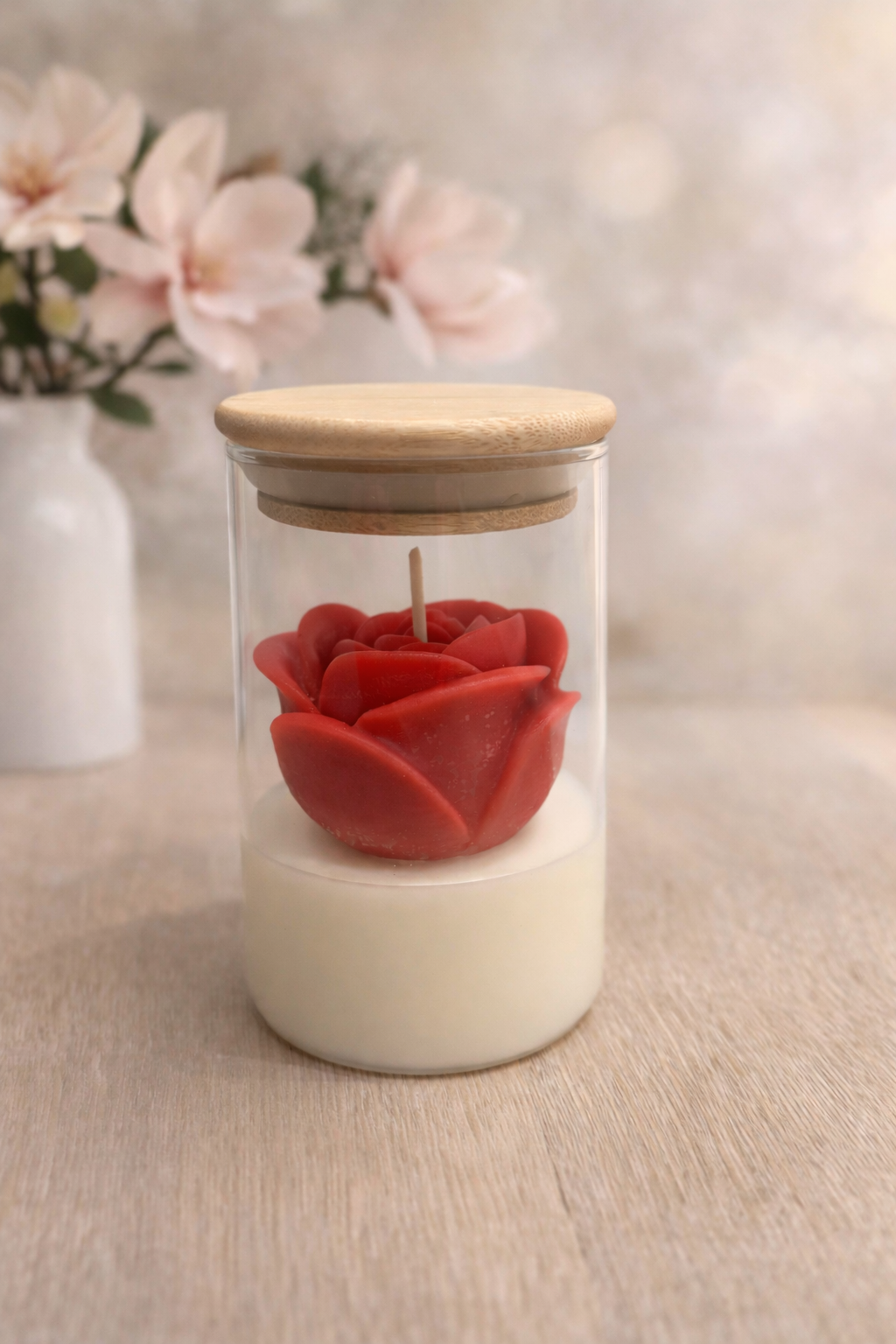 Een kaars in de vorm van een rode roos in een glazen pot met houten deksel, met bloemen op de achtergrond.