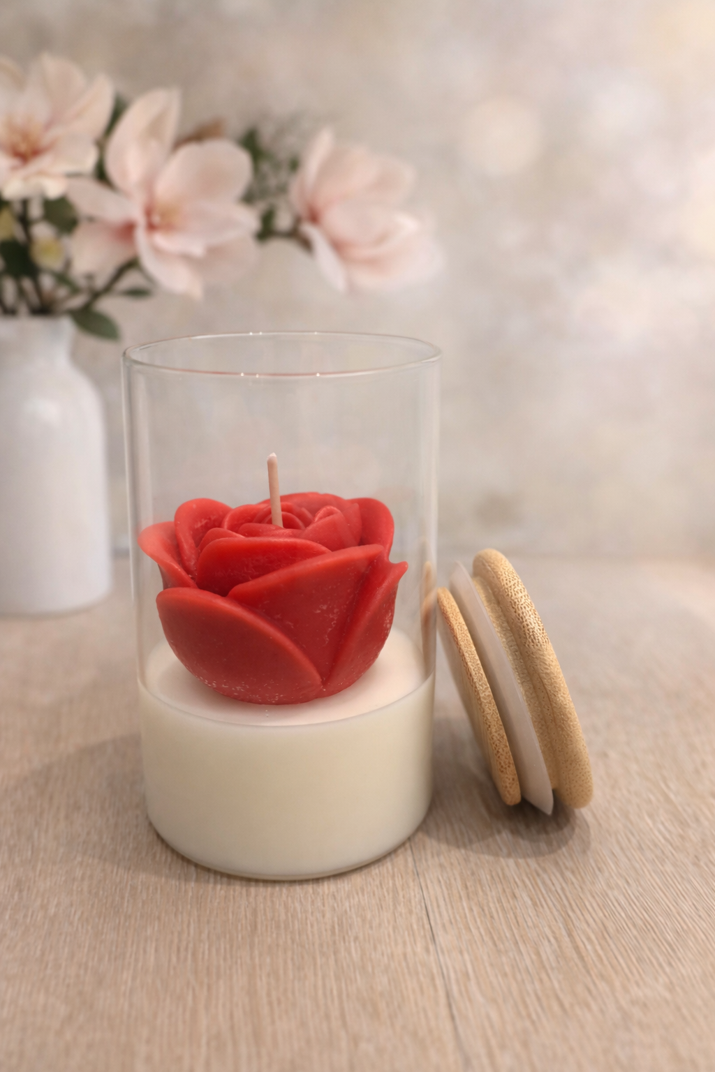 Een kaars in de vorm van een rode roos in een glazen pot met houten deksel, met bloemen op de achtergrond.