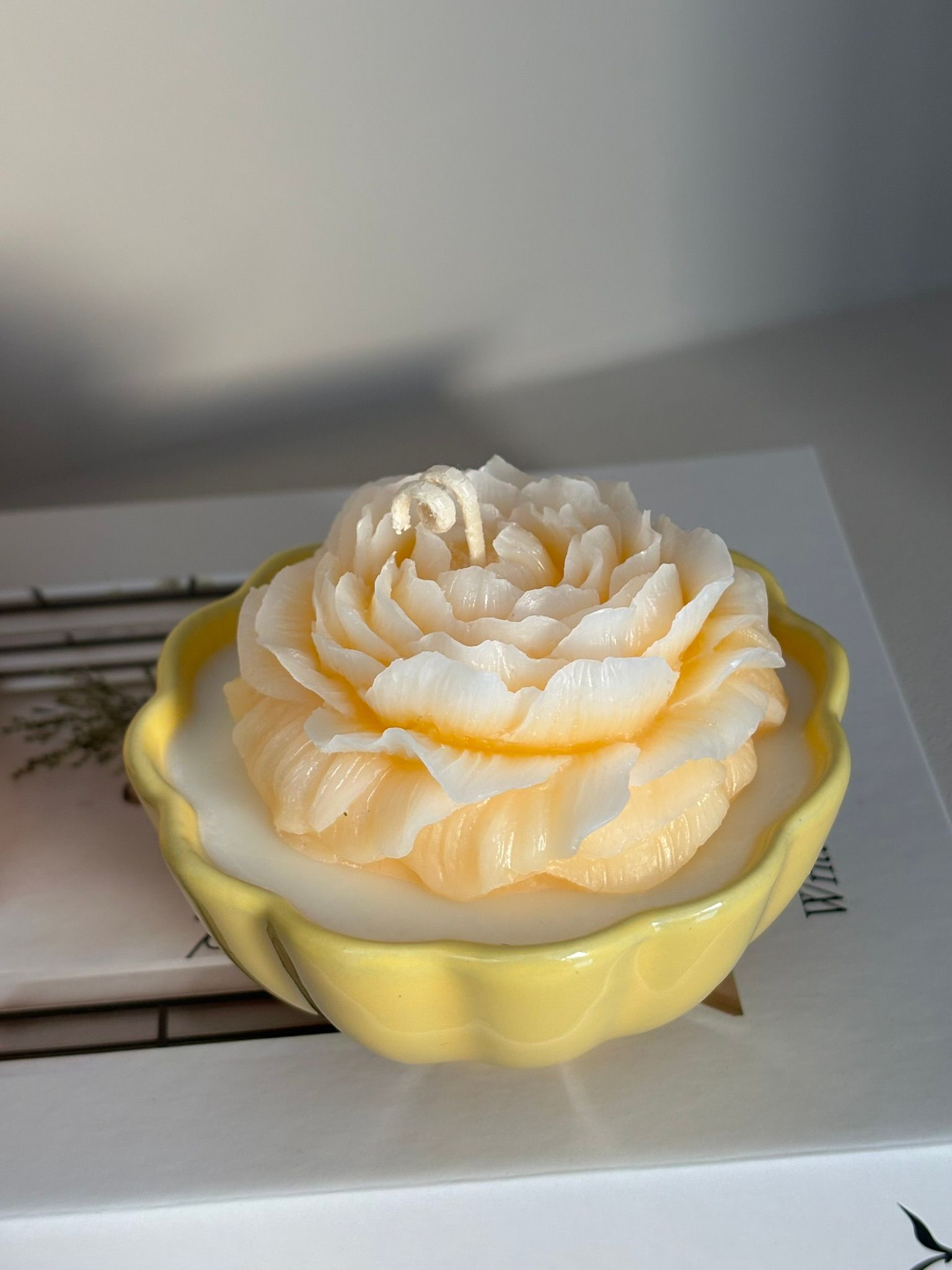 Bloemvormige kaars met gele en witte bloemblaadjes, in een geel gegolfd schaaltje, op een witte ondergrond.