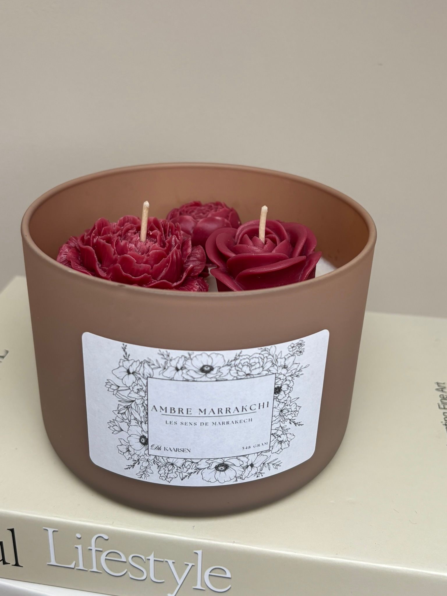 Een kaars in een bruine houder met daarin drie rode, bloemvormige kaarsen, op een stapel boeken.