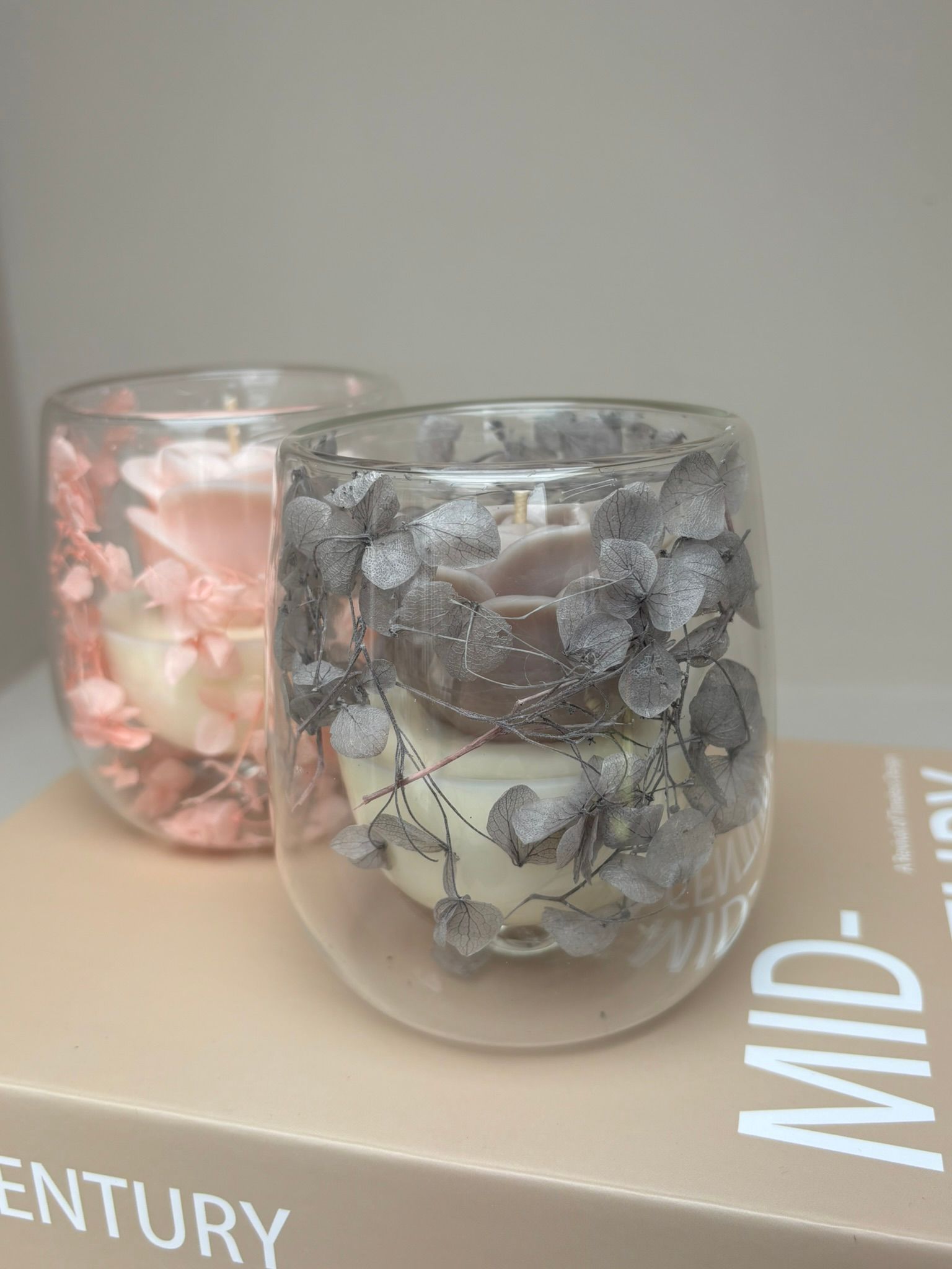 Twee glazen kaarsenstandaards, een roze en een grijze, met bloemversieringen, op een boek.