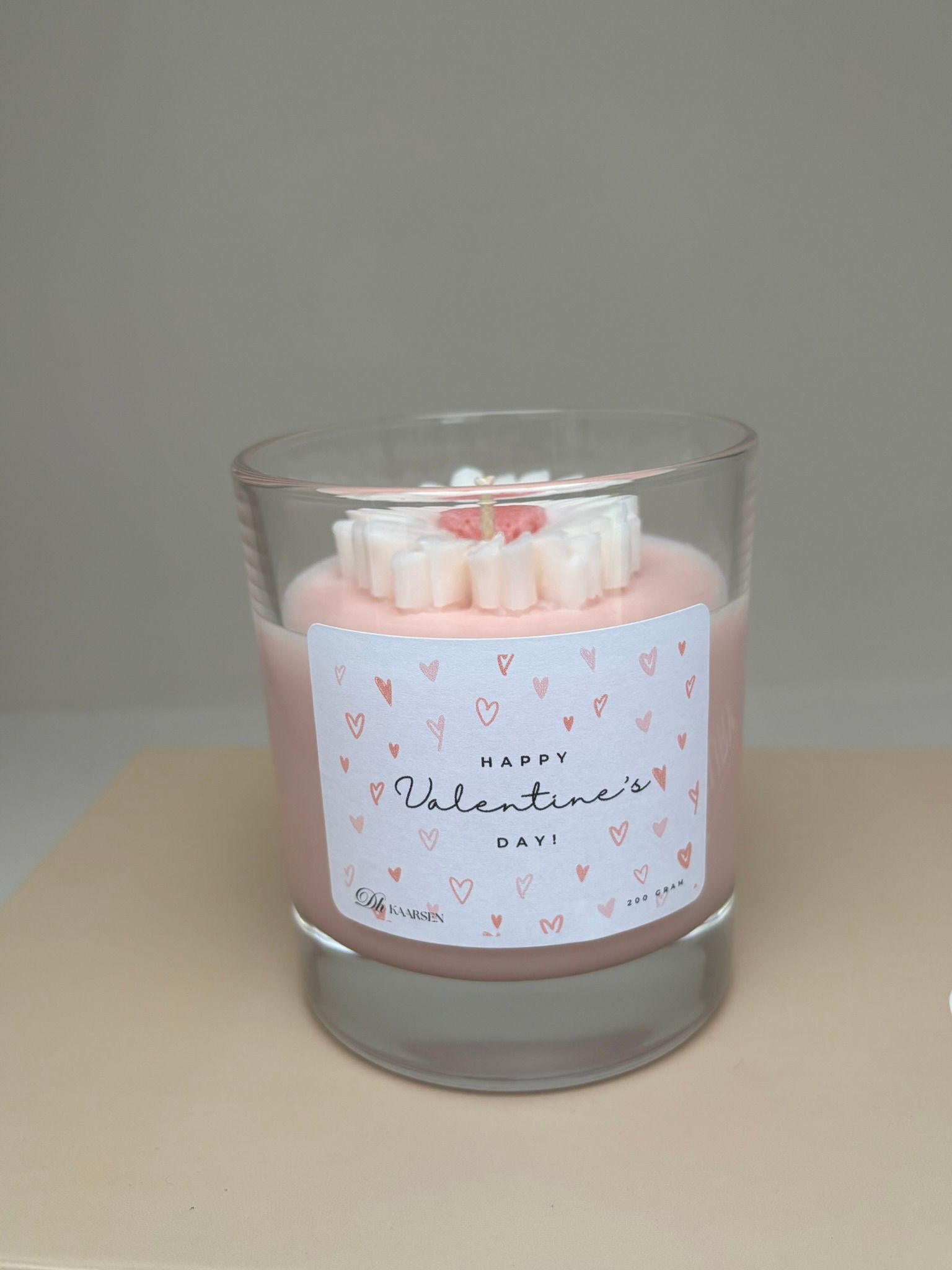 Roze kaars in een helder glazen potje met een hartvormig label en een bloemversiering bovenop.