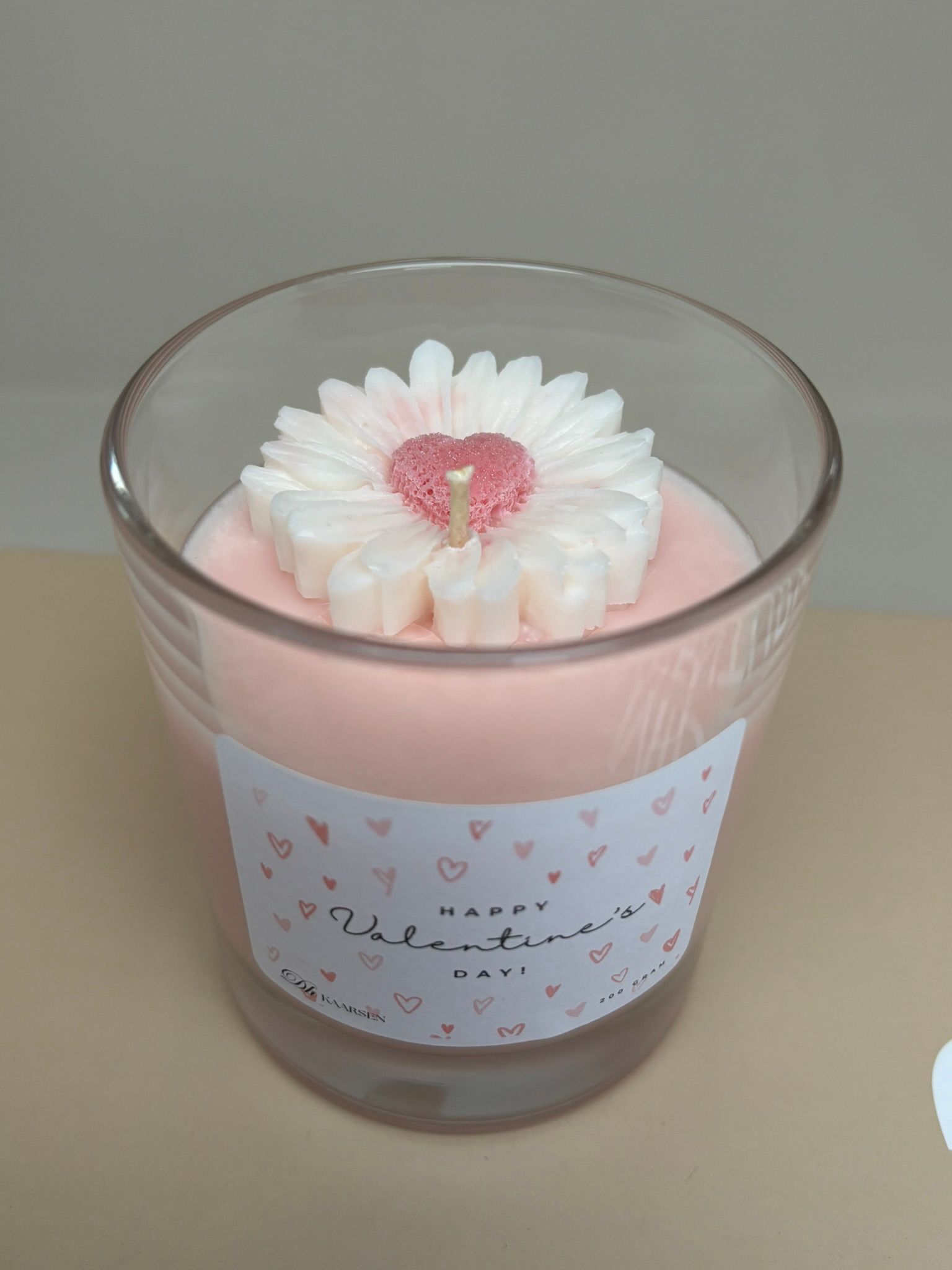 Roze kaars in een helder glazen potje met een hartvormig label en een bloemversiering bovenop.