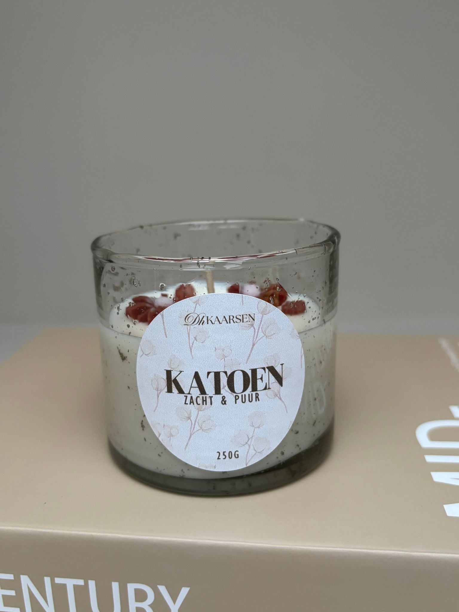 Een kaars in een glazen pot met etiket, die op een lichtgekleurd boek rust, tegen een effen achtergrond.