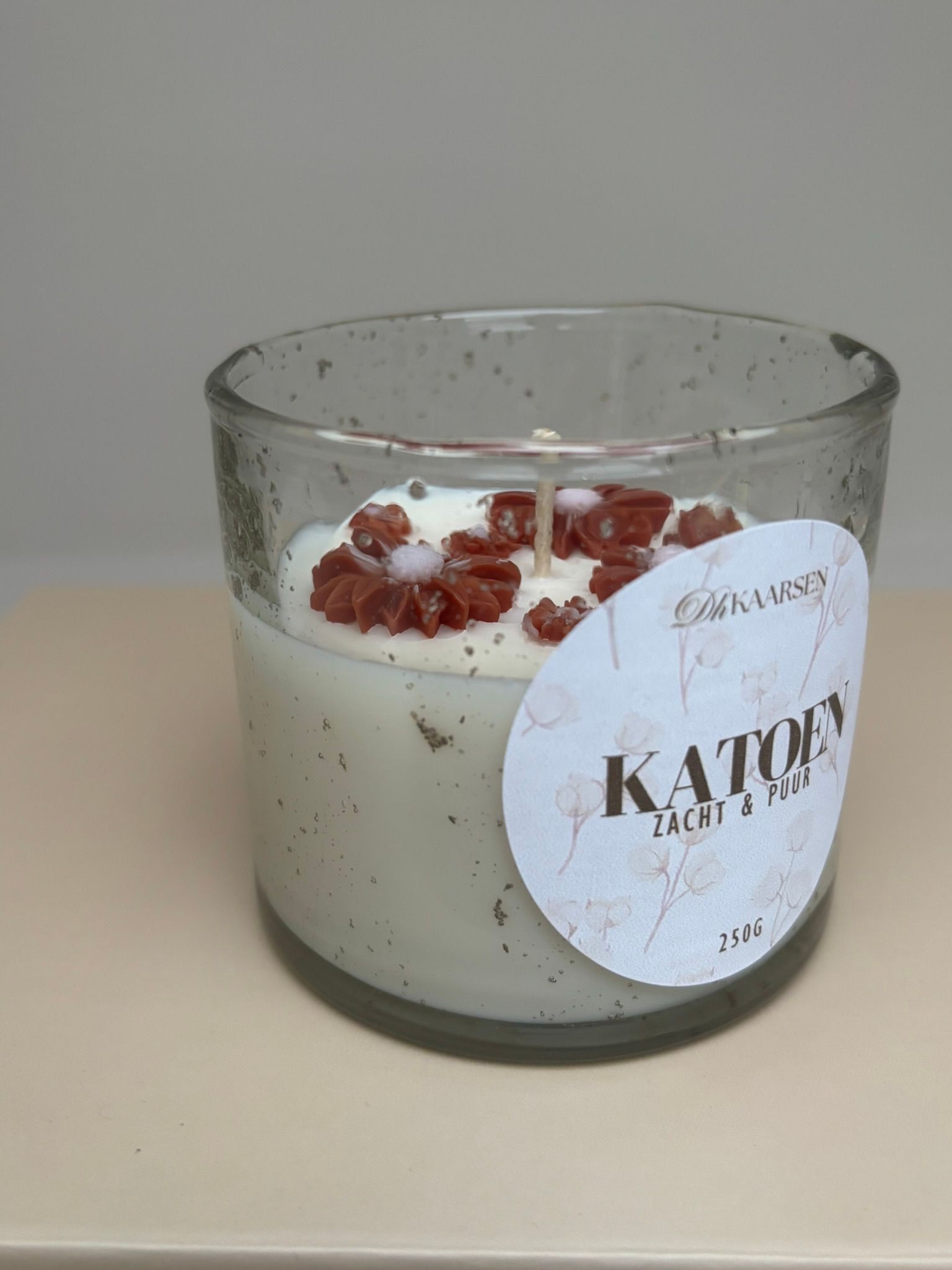 Een kaars in een glazen pot met etiket, die op een lichtgekleurd boek rust, tegen een effen achtergrond.