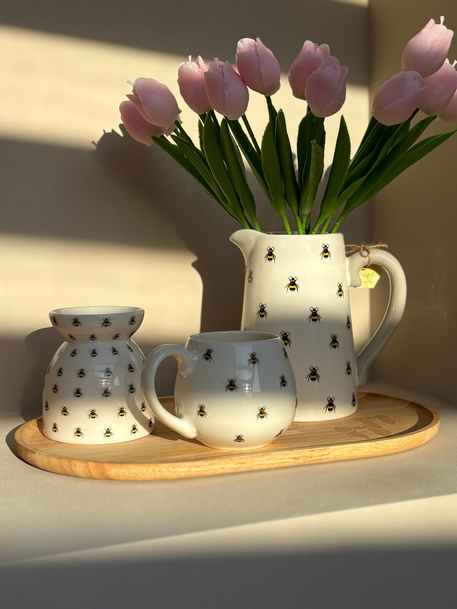 Een witte keramische waxbrander, versierd met kleine bijenmotieven, staat op een rond houten dienblad in het zachte zonlicht.