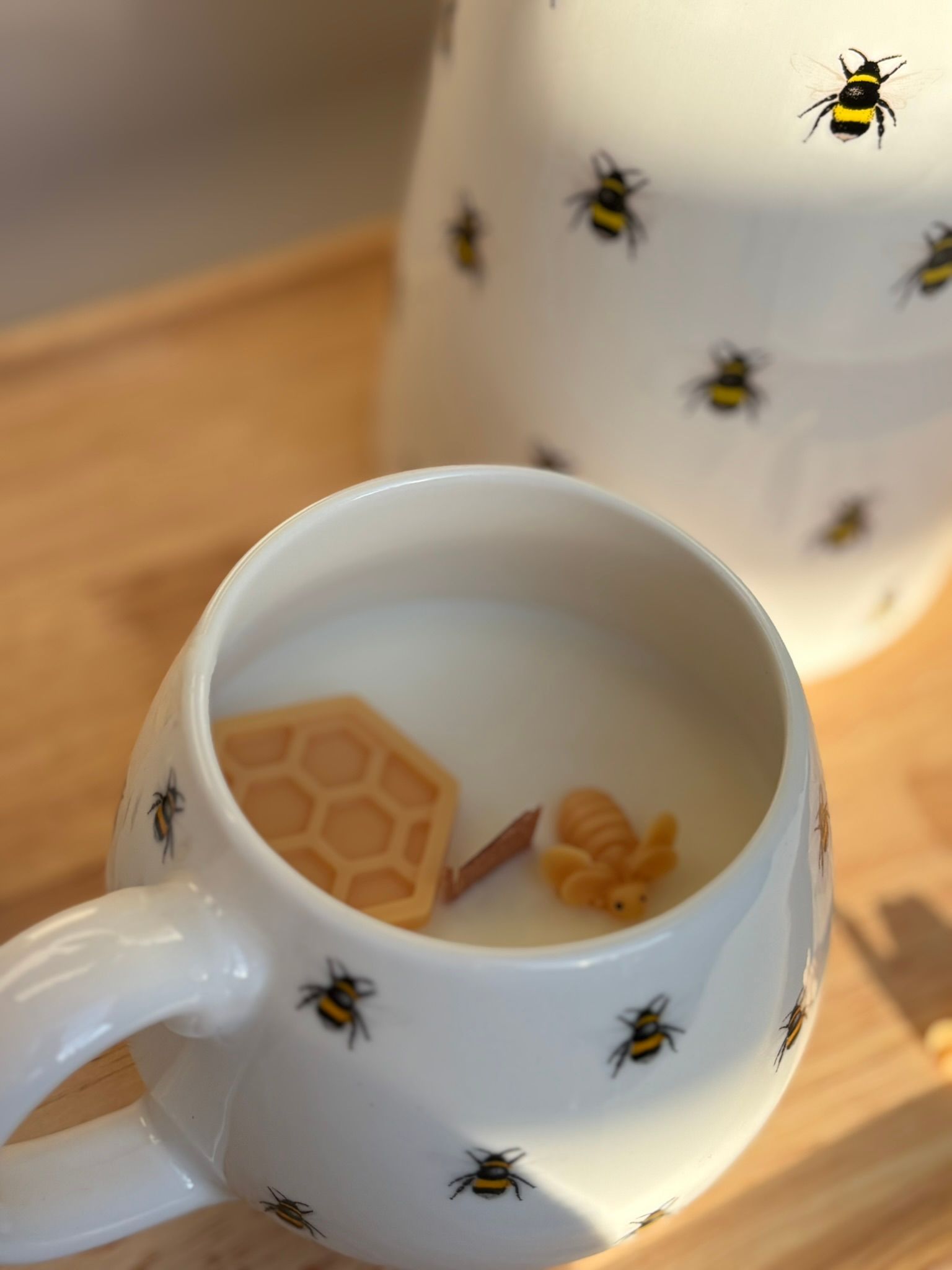 Een witte mok en bijpassende kan versierd met bijenmotieven, met een honingraatvormige kaars in de mok.