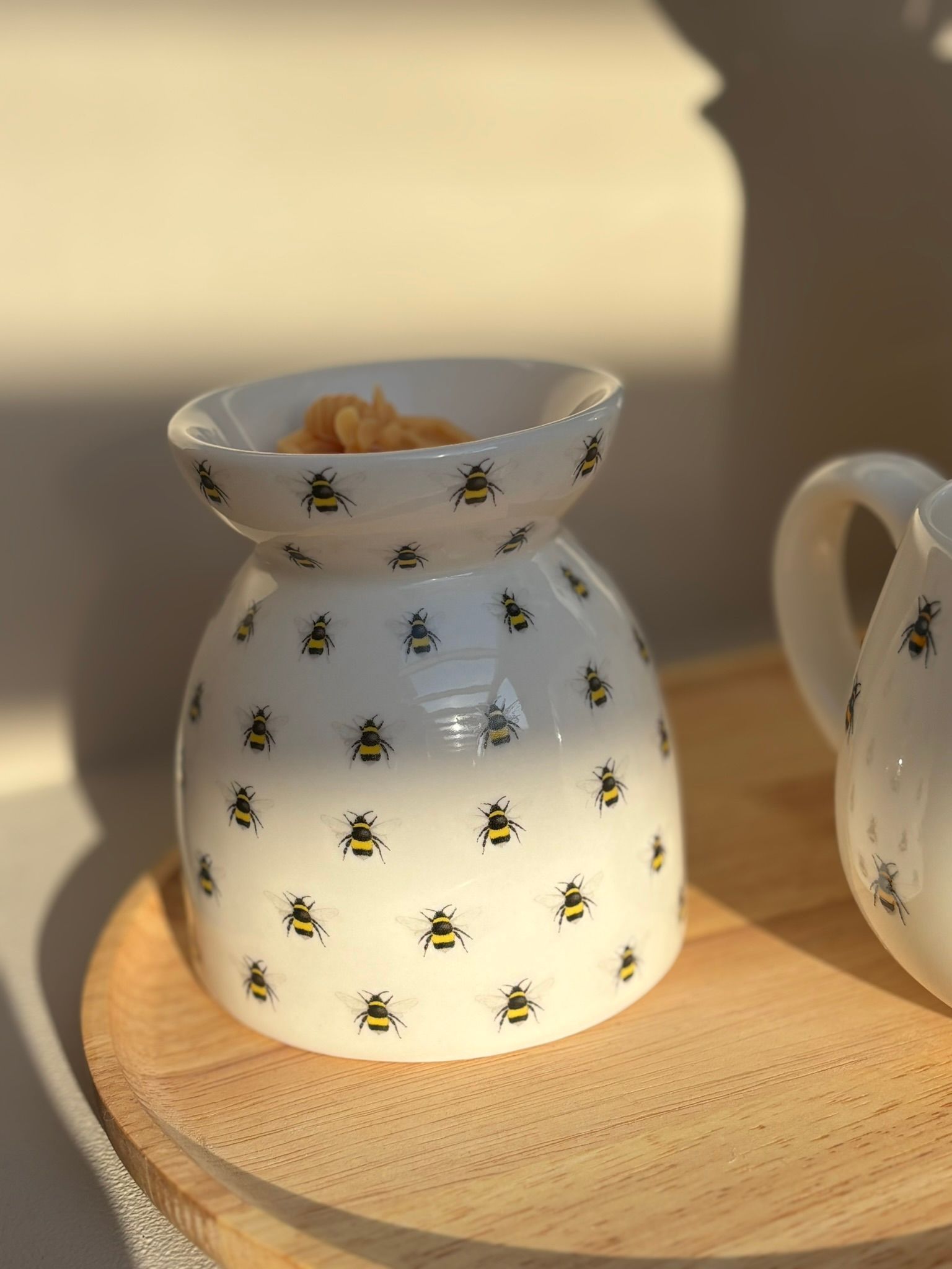 Een witte keramische waxbrander, versierd met kleine bijenmotieven, staat op een rond houten dienblad in het zachte zonlicht.
