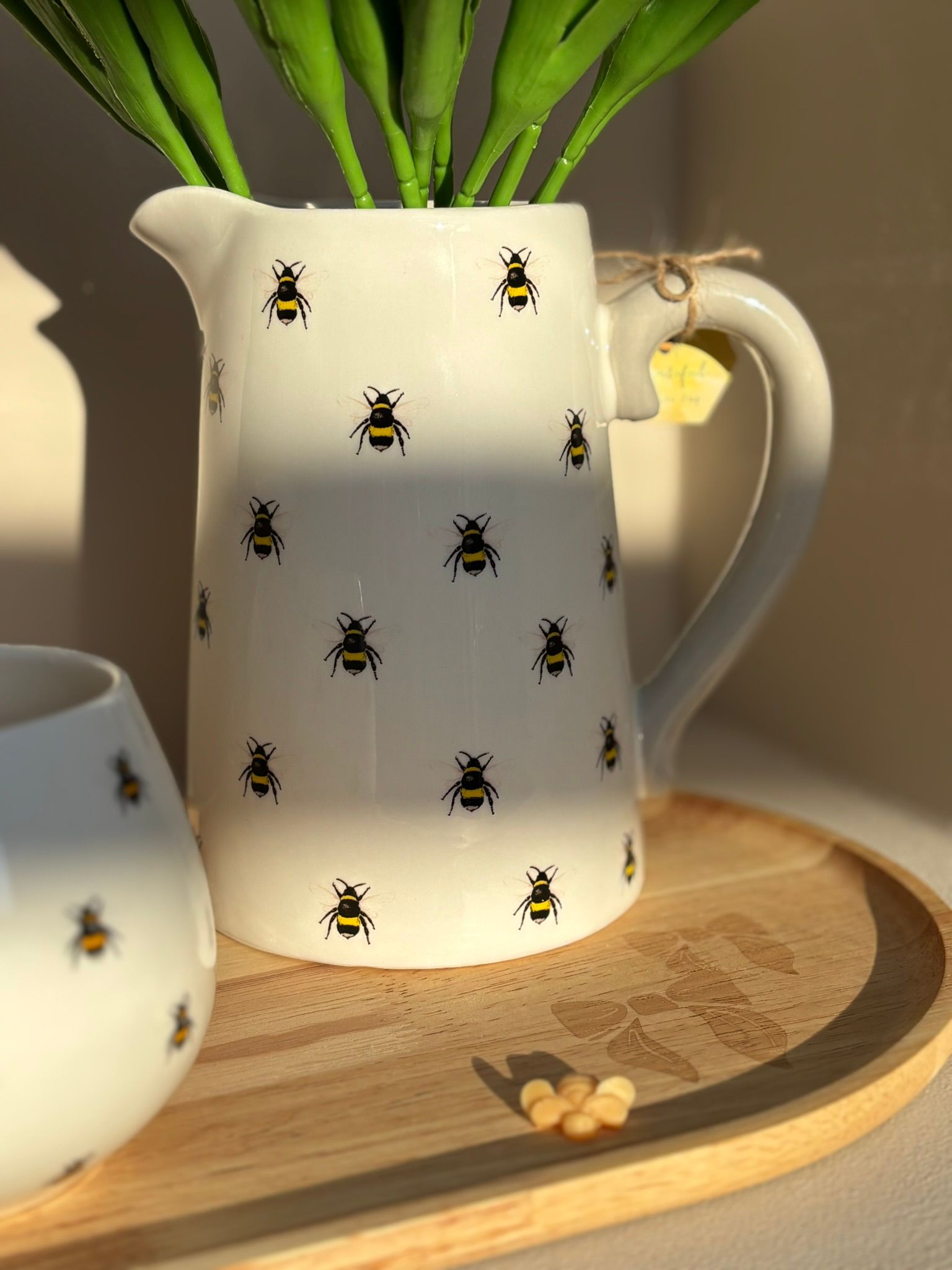 Een kan, een mok en een klein potje met bijenmotieven op een houten dienblad, gevuld met roze tulpen in zacht zonlicht.