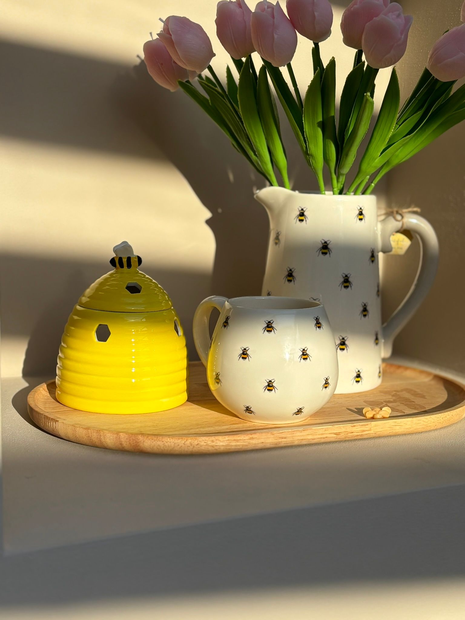 Een gele bijenkorf, een kan met bijenmotief en roze tulpen, en een bijpassend klein melkkannetje op een houten dienblad.