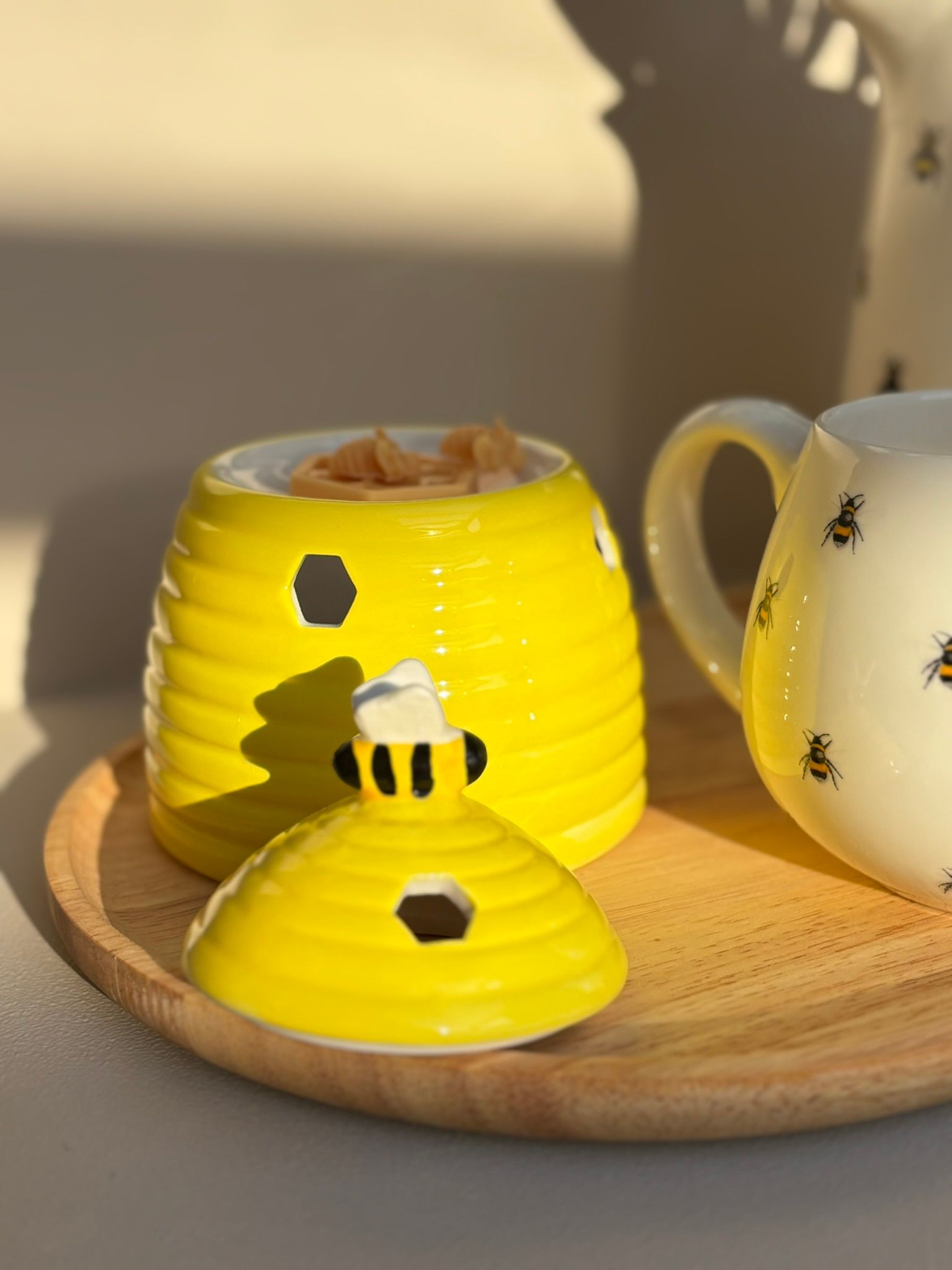 Een gele bijenkorf, een kan met bijenmotief en roze tulpen, en een bijpassend klein melkkannetje op een houten dienblad.