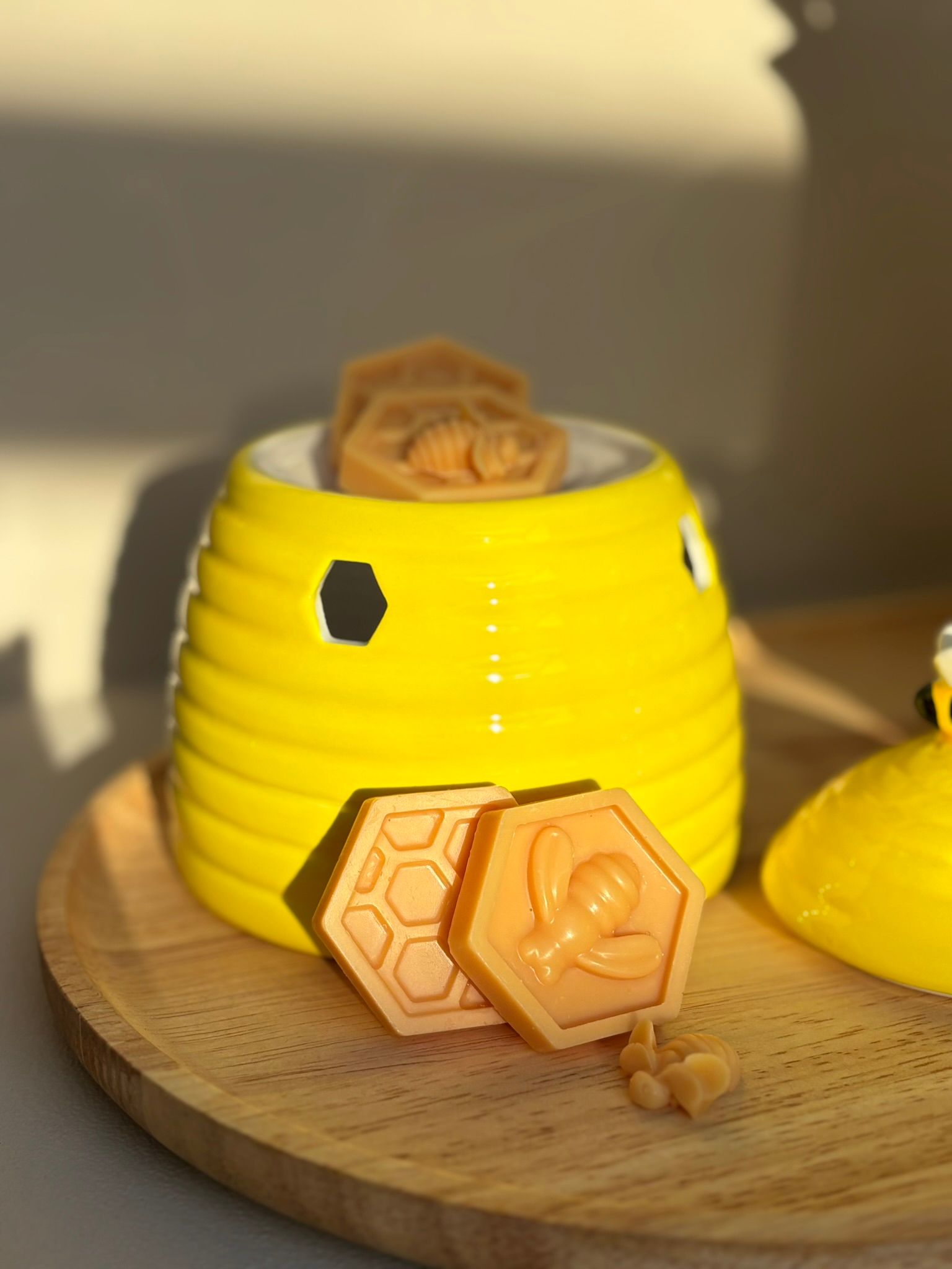 Een felgele, bijenkorfvormige waxbrander op een houten dienblad, met honingraat- en bijenvormige waxblokjes ervoor.