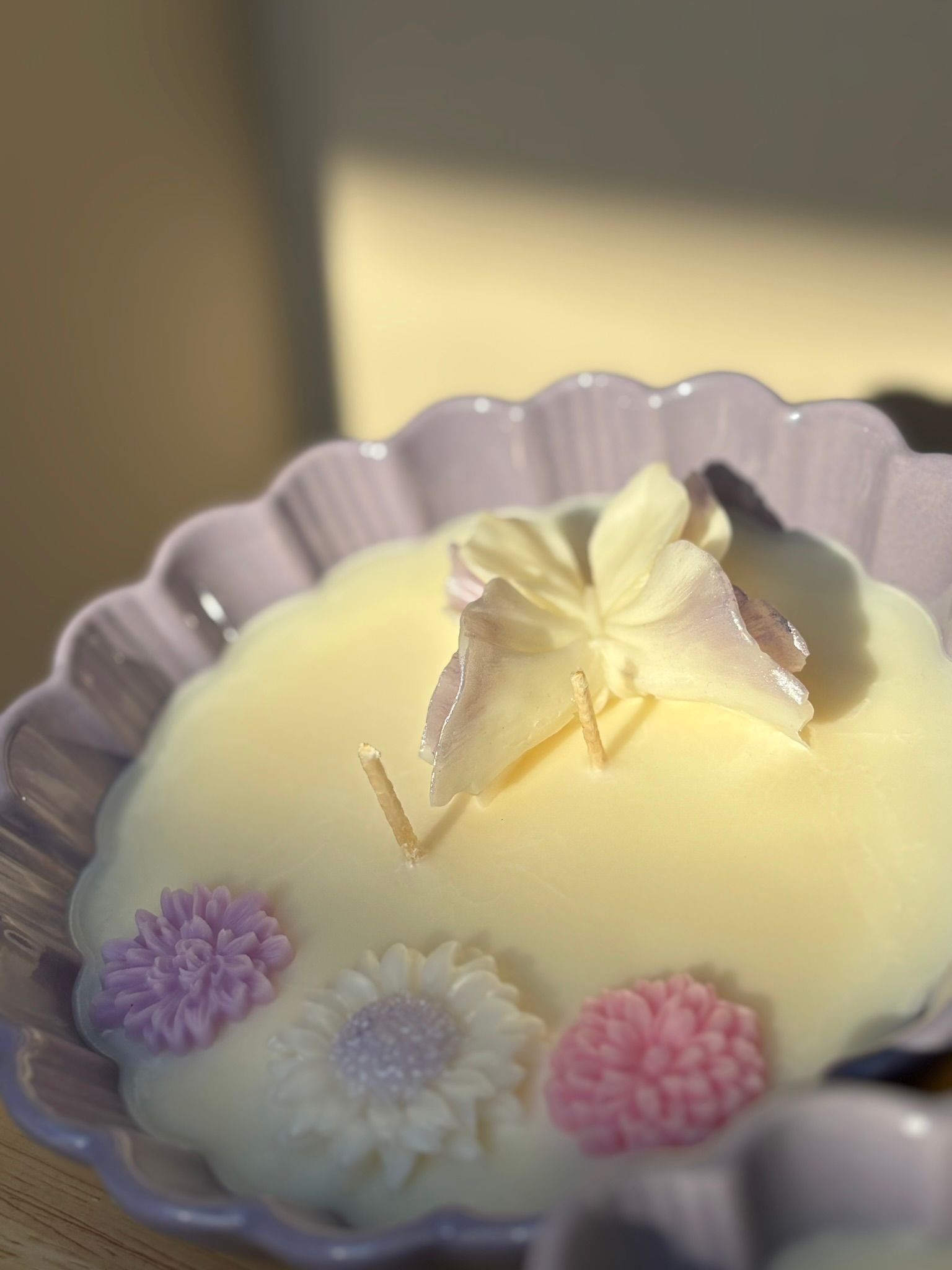 Een kaars in een gegolfde lavendelkleurige schaal naast een vaas met bijenmotief en groene stelen op een houten dienblad.