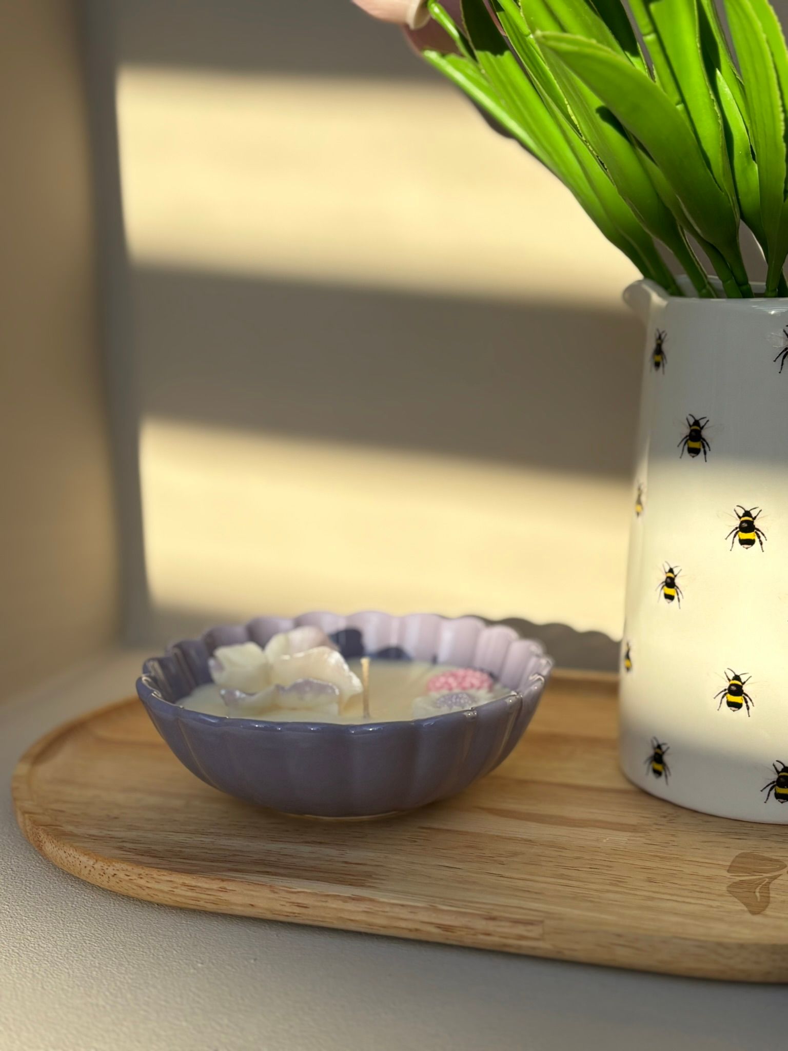 Een kaars in een gegolfde lavendelkleurige schaal naast een vaas met bijenmotief en groene stelen op een houten dienblad.