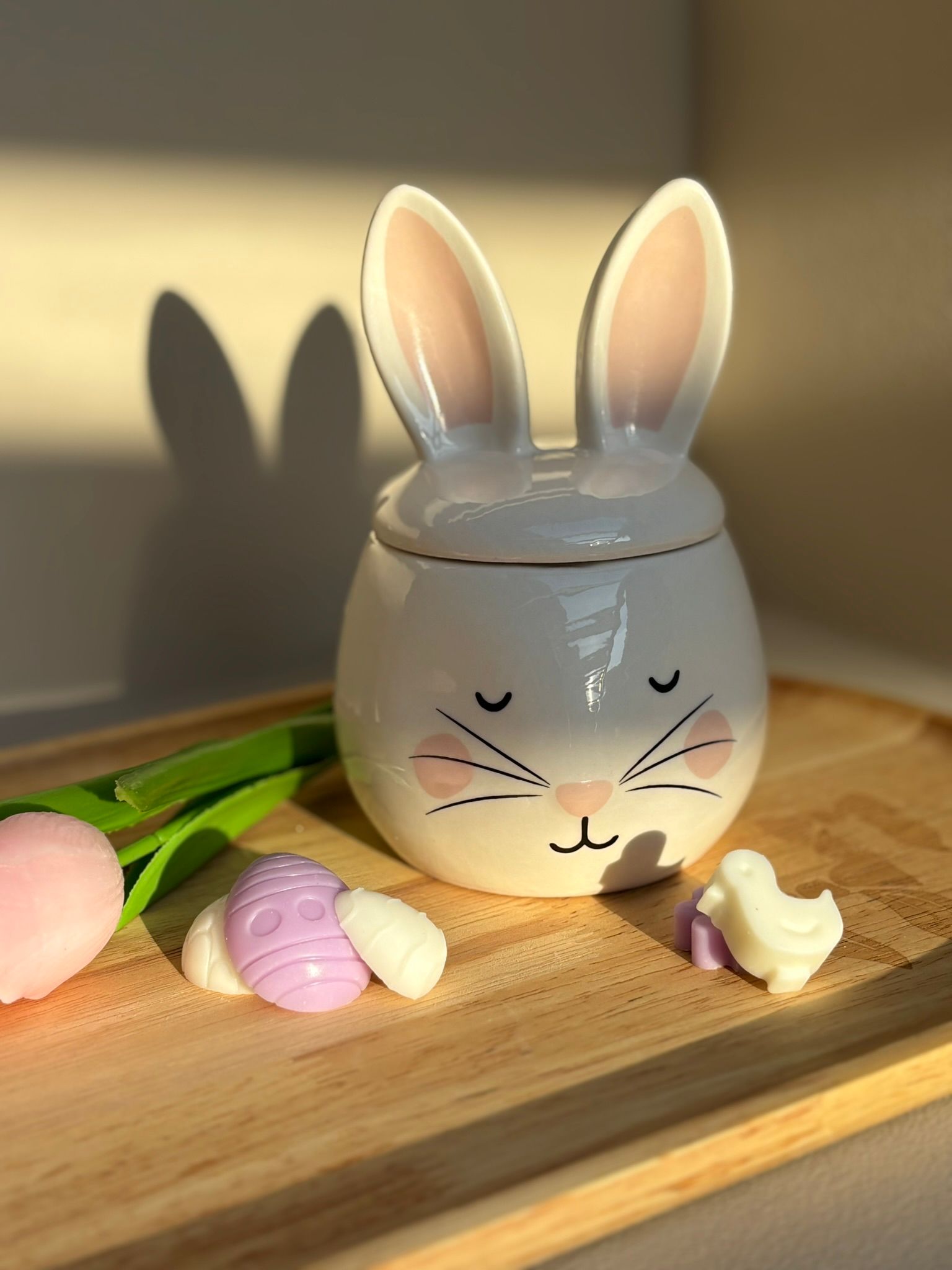 Een keramische konijnenpot met gesloten ogen en roze wangen, staand op een houten dienblad met twee kleine paasornamenten.