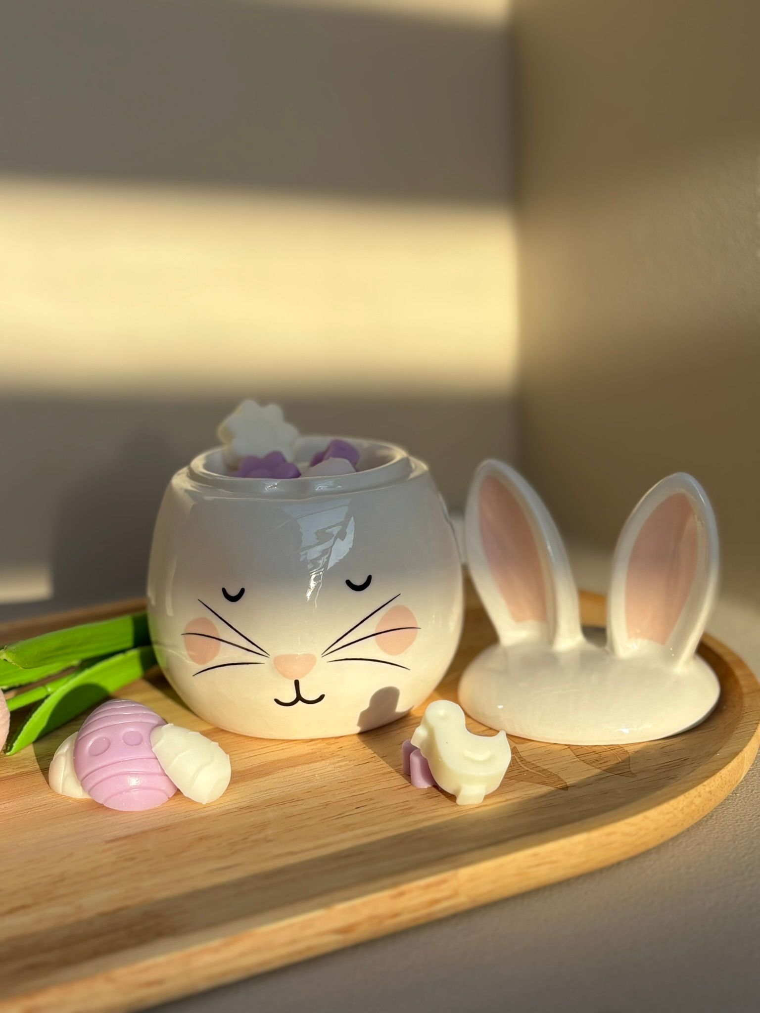 Een keramische konijnenpot met gesloten ogen en roze wangen, staand op een houten dienblad met twee kleine paasornamenten.