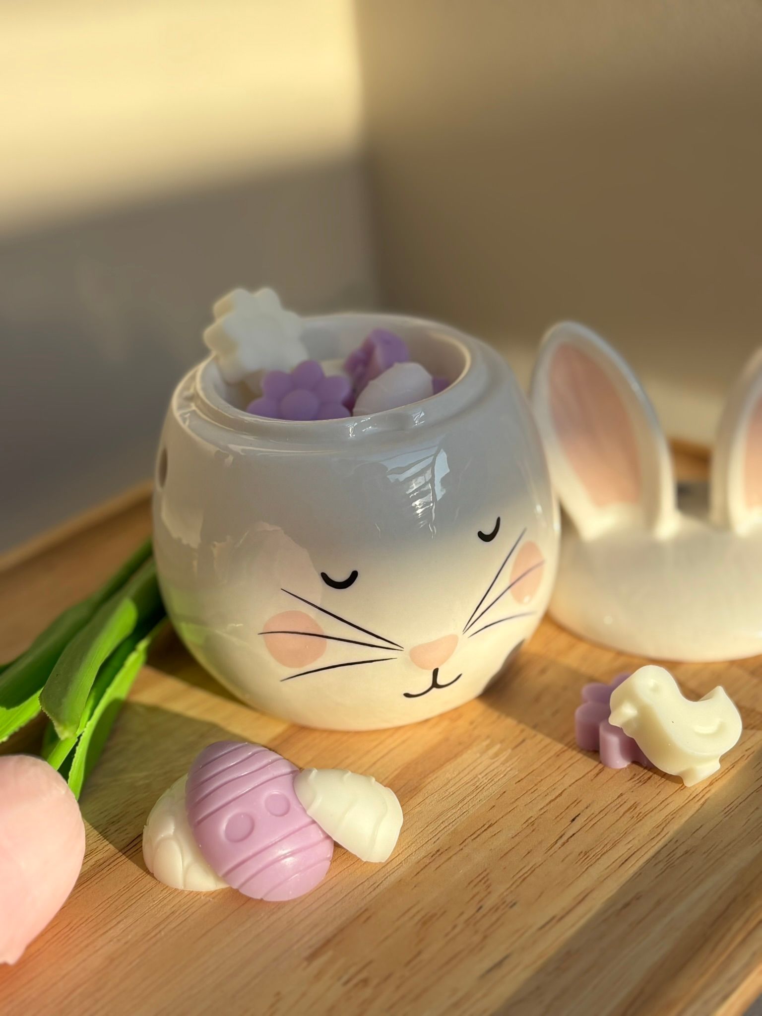 Een witte keramische konijnenkom gevuld met kleine, pastelkleurige wasvormpjes, die op een houten dienblad met decoratieve voorwerpen staat.