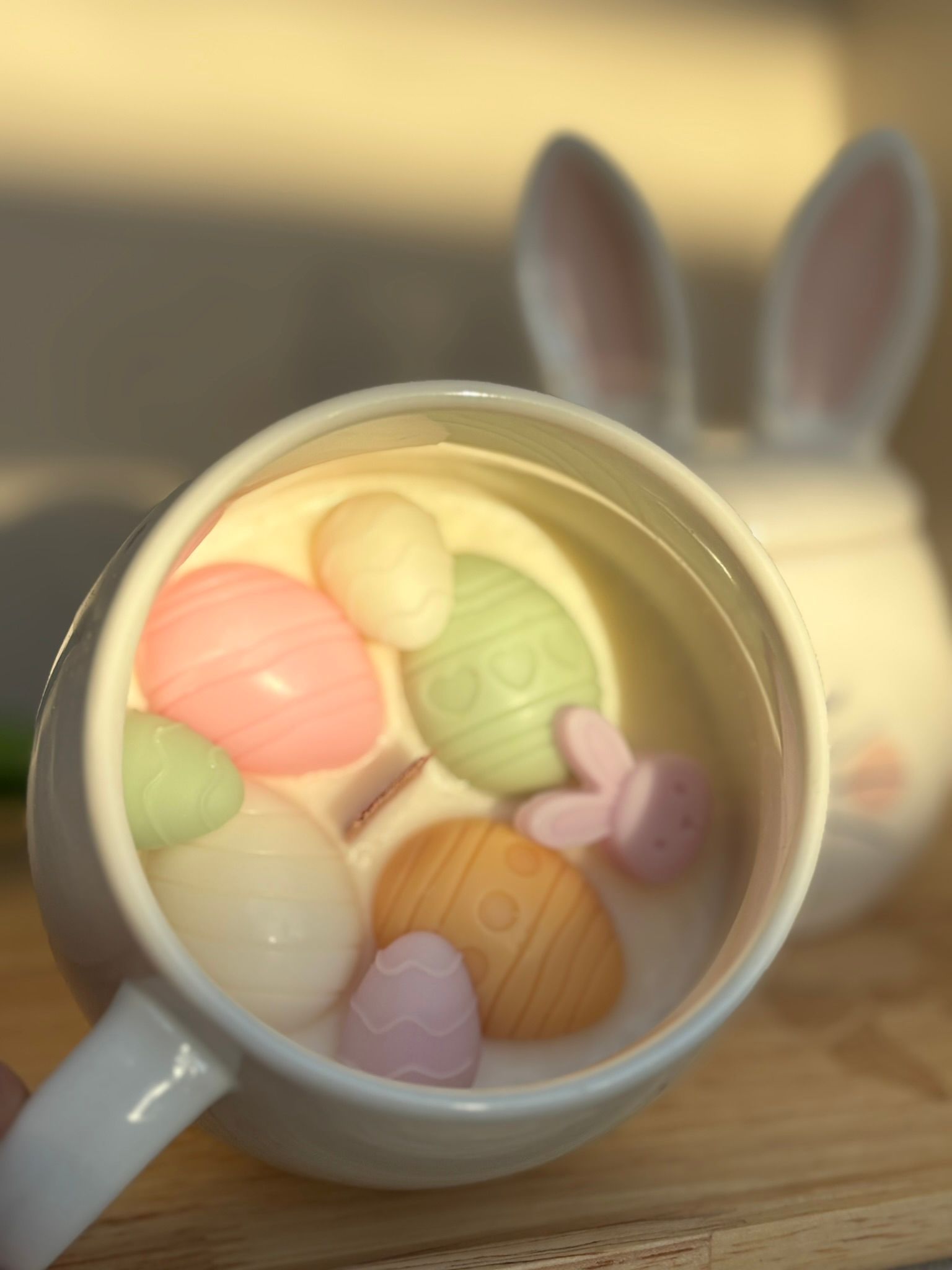 Een witte keramische mok met een geschilderde afbeelding van een konijntje met een roze bloemenkrans, zittend op een houten ondergrond.