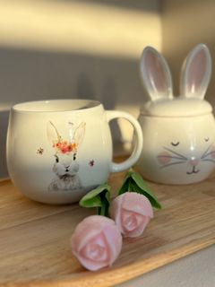 Een keramische mok in de vorm van een konijn en een potje met deksel in de vorm van een konijn, versierd met twee roze bloemetjes, op een houten dienblad.