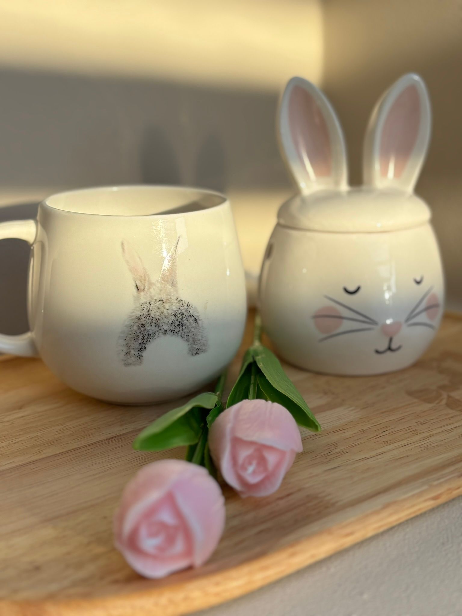 Een mok met een konijntje met een bloemenkroon, een potje met een konijntje erop en twee lichtroze bloemknopjes op een houten dienblad.